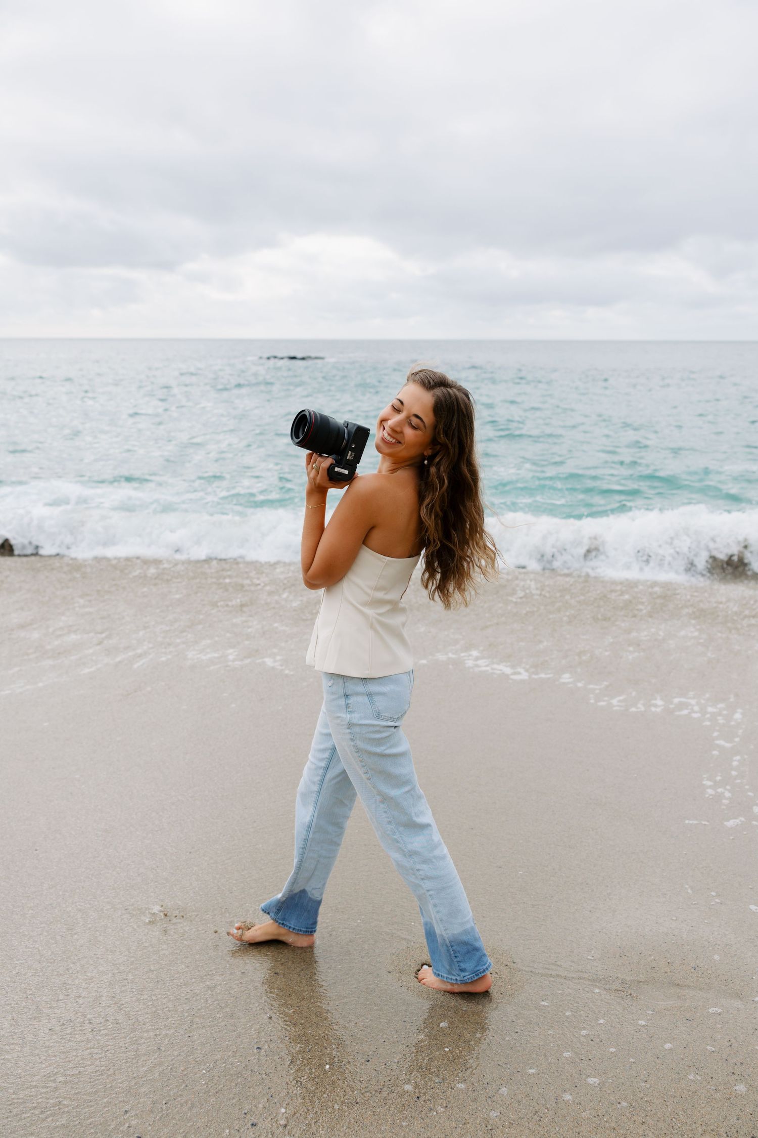 Photographer with long hair wearing white top and jeans captures coastal scenery on beach with waves crashing behind.