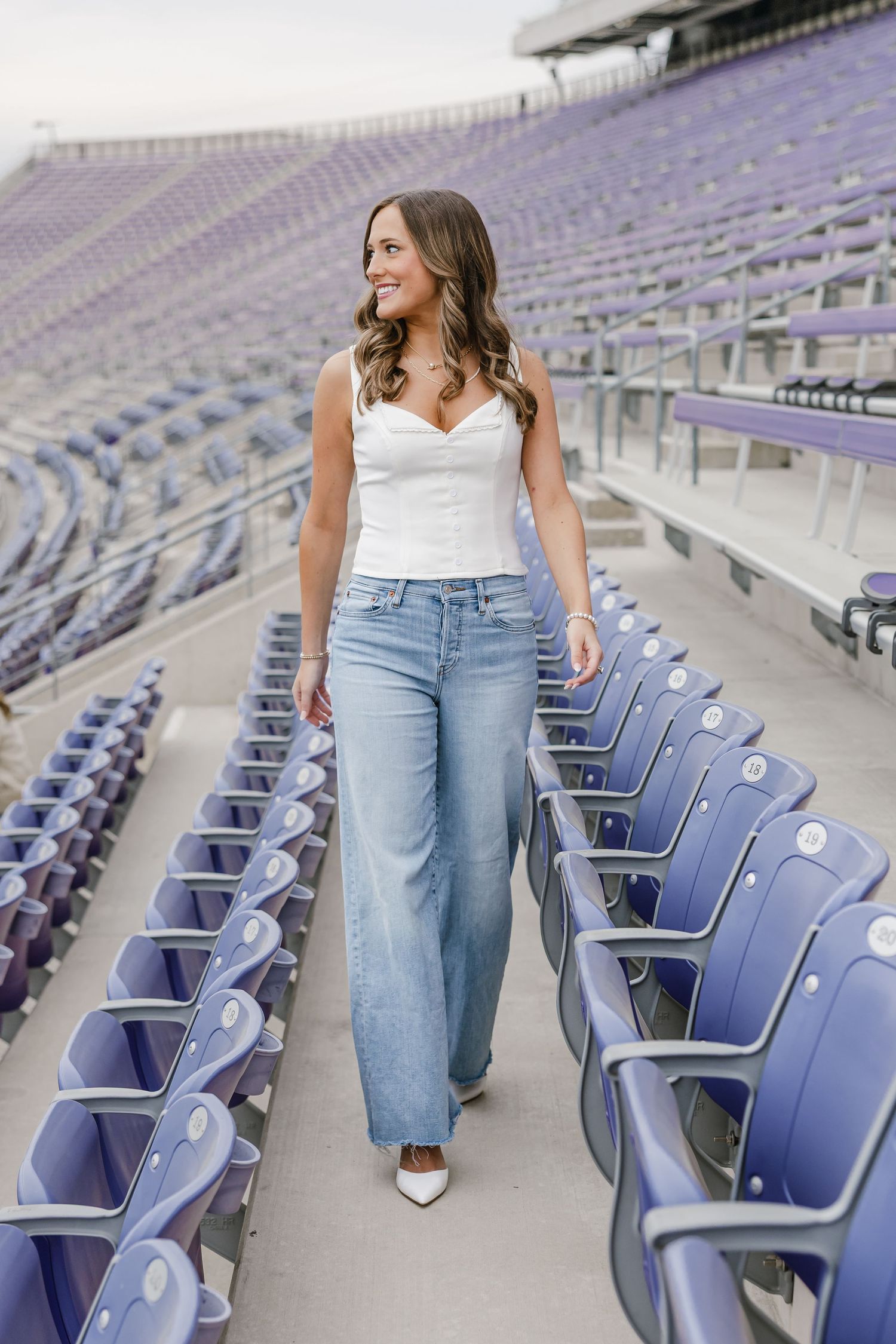 Woman in white corset top and wide-leg jeans walking through purple stadium bleachers, smiling confidently.