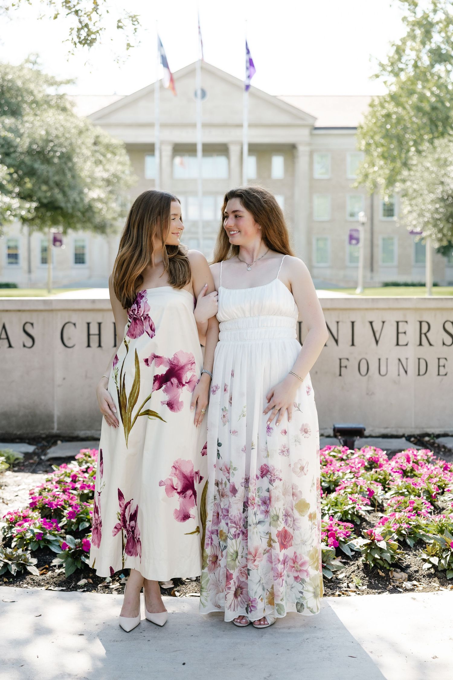 Two women in floral maxi dresses pose together outside Texas Christian University on a sunny day.