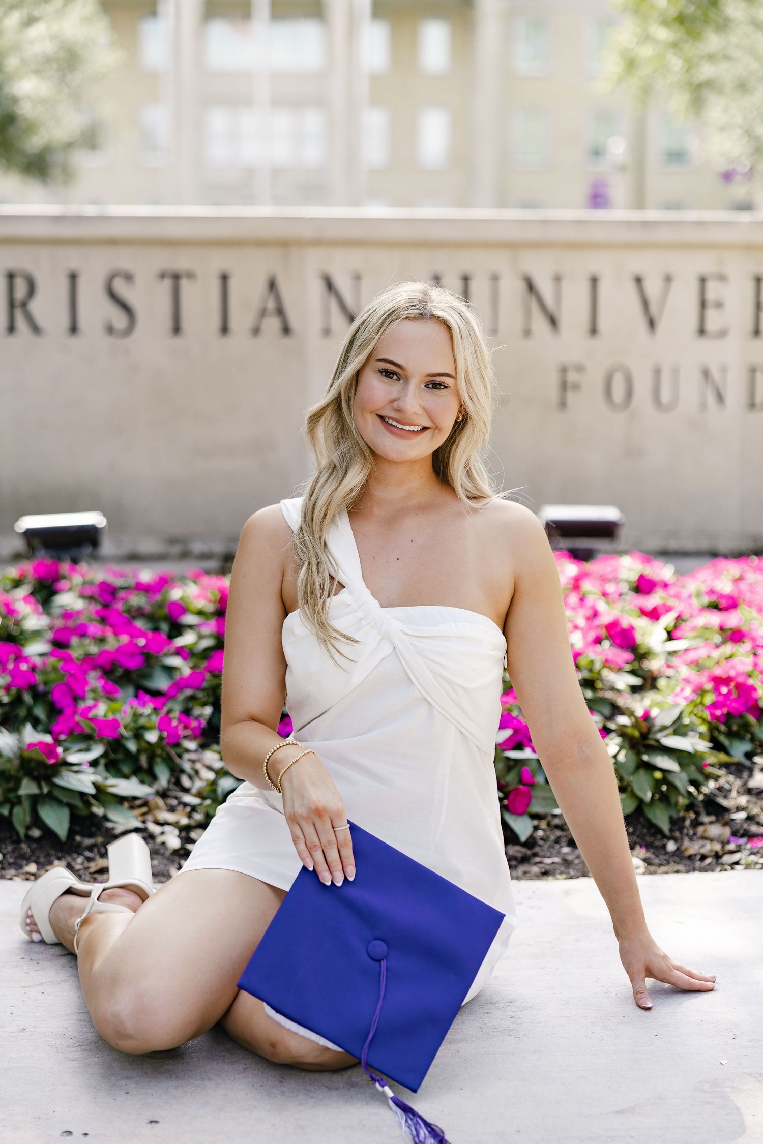 Smiling blonde graduate in white dress holds blue cap at Christian University Foundation sign.