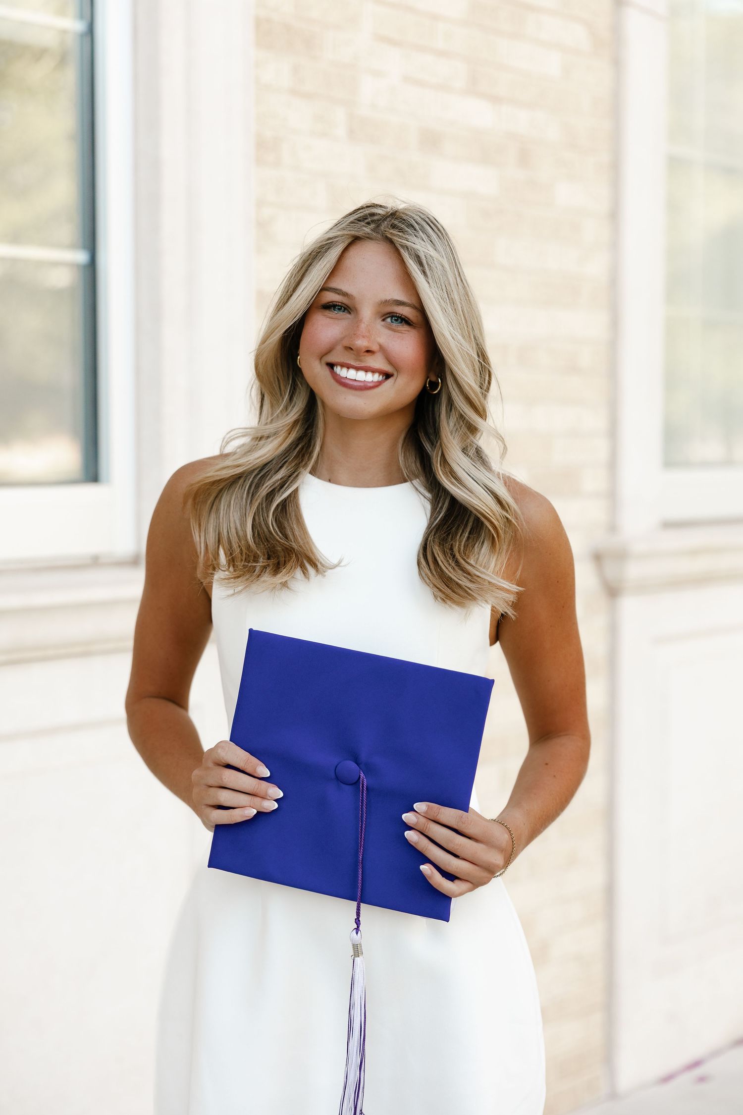 Smiling young woman in white dress holding blue graduation cap outdoors.