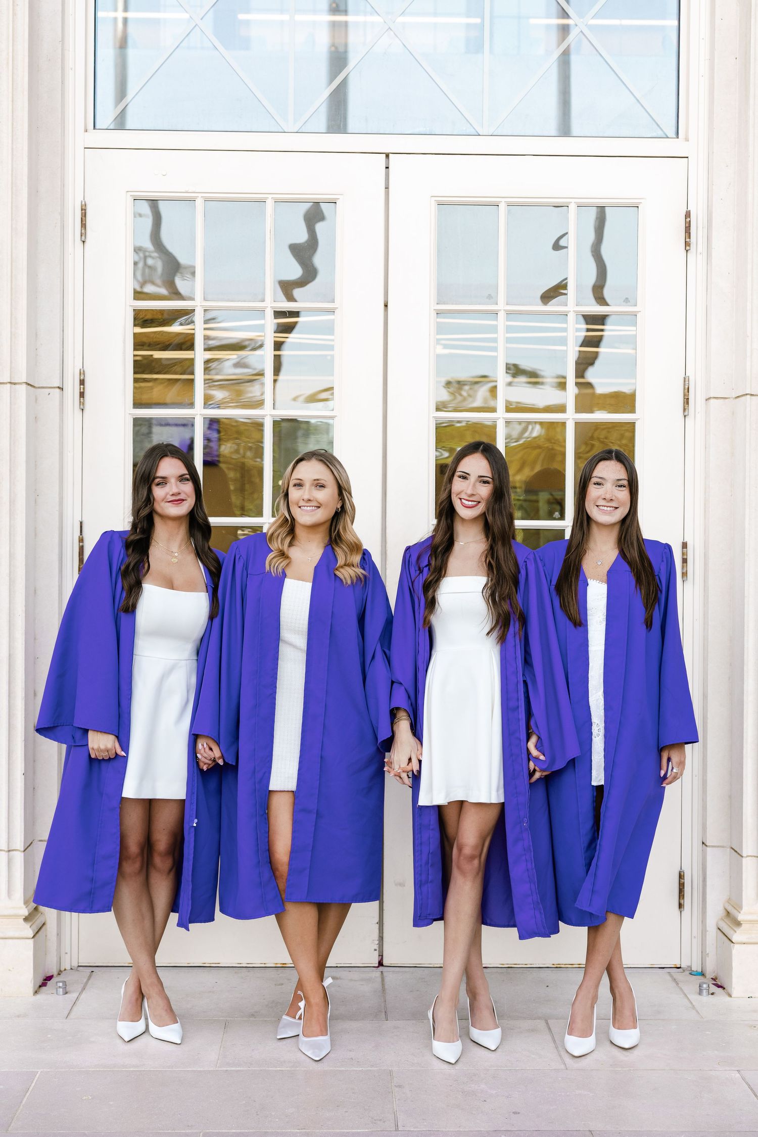 Four female graduates in royal blue graduation gowns and white dresses pose together in front of white doors.