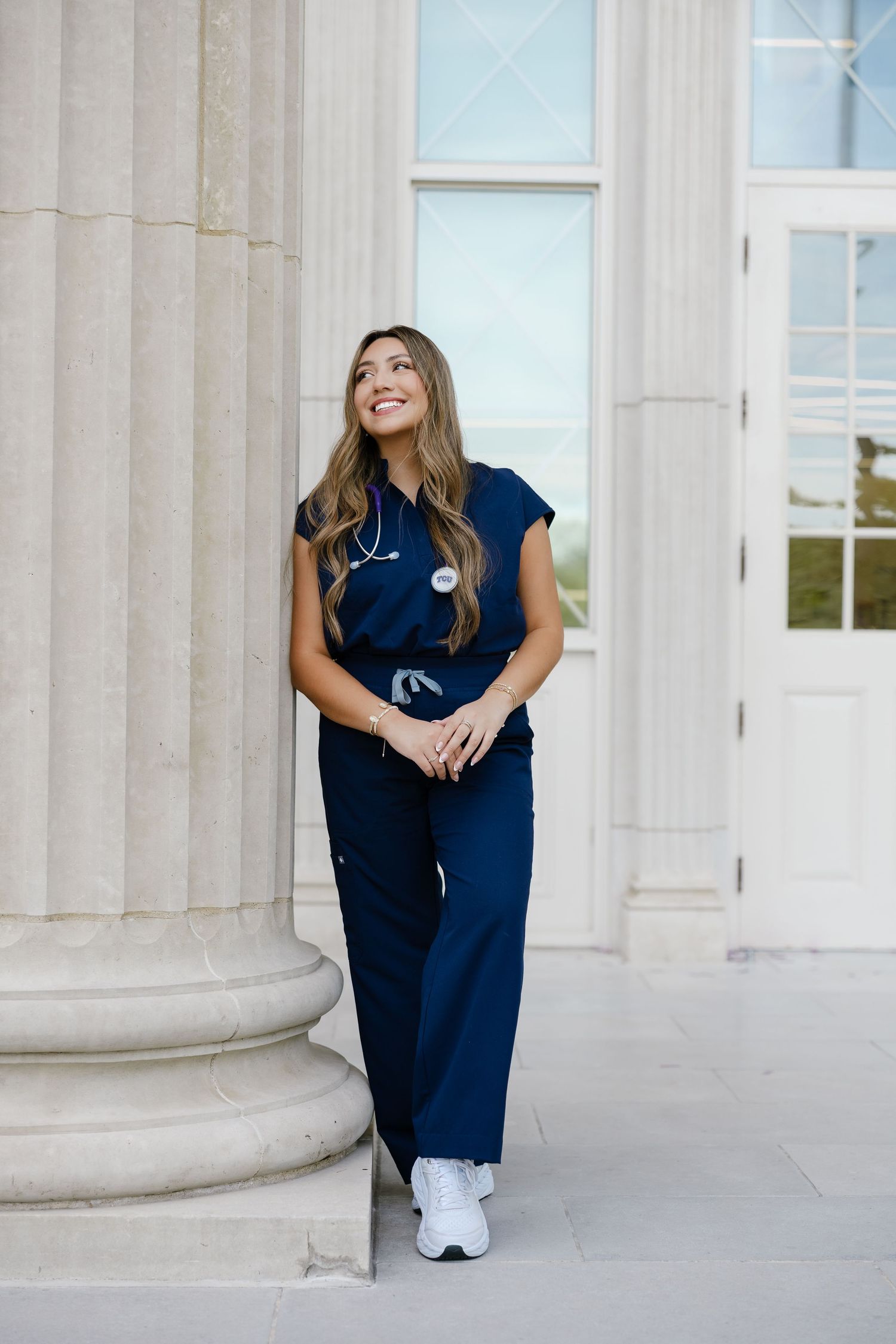 Smiling nursing student in navy scrubs with stethoscope leaning against a column outside a building at Chapman University
