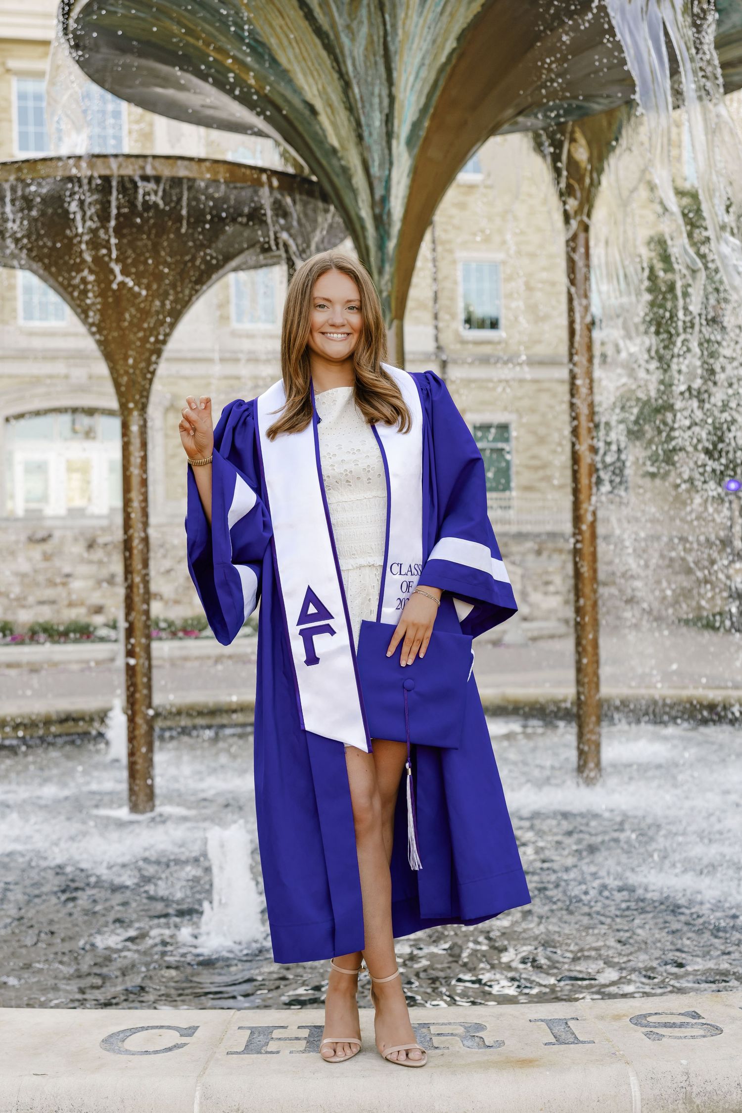Young woman in blue graduation gown and white stole with Greek letters poses proudly in front of a decorative fountain.