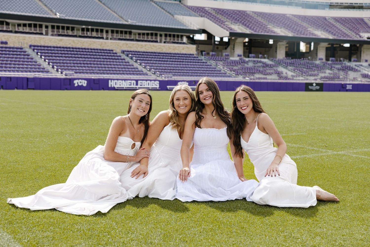 Four women in white dresses pose together on a green football field at a purple-themed stadium.