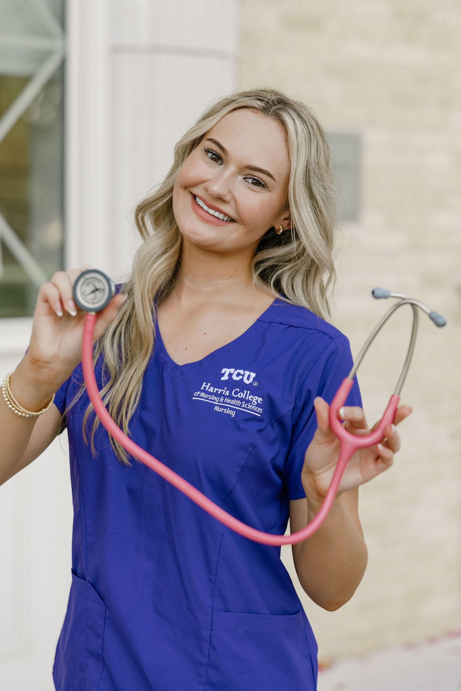 Smiling nursing student in TCU Harris College purple scrubs holding a pink stethoscope outdoors.