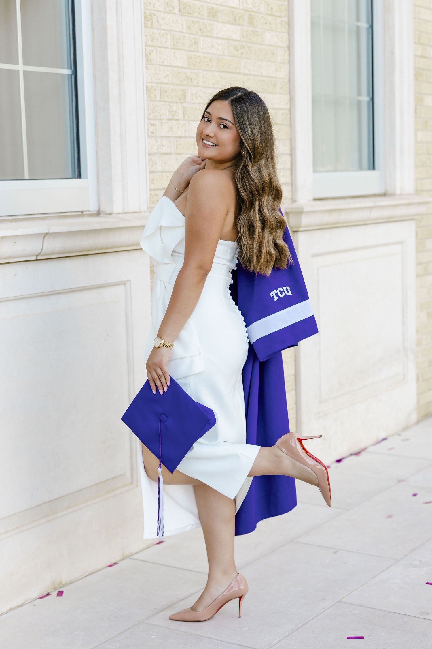 A smiling young woman in a white dress holds a TCU graduation cap and gown while posing outdoors in nude heels.