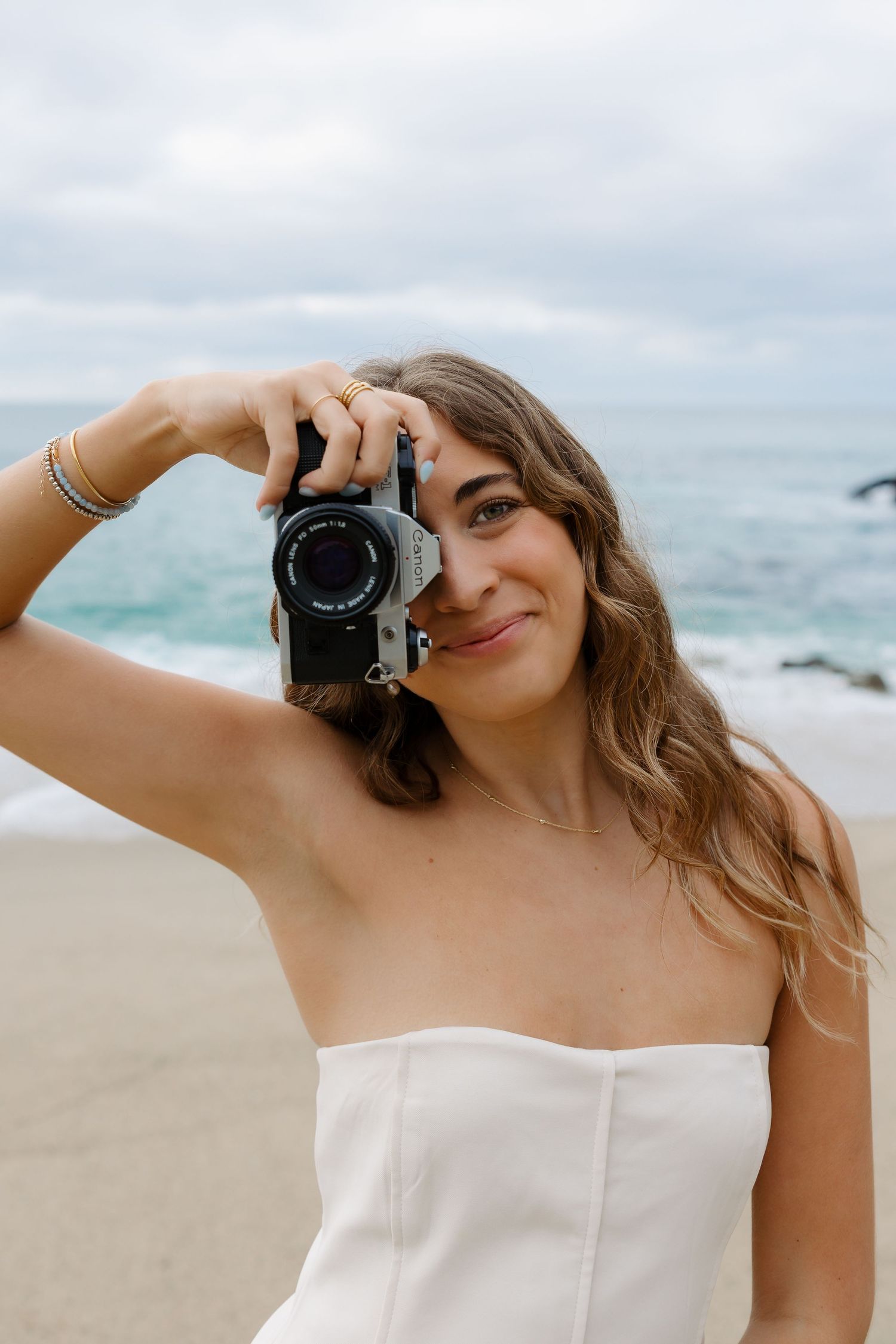 A photographer in a white dress takes self portraits on a cloudy beach with waves in the background.