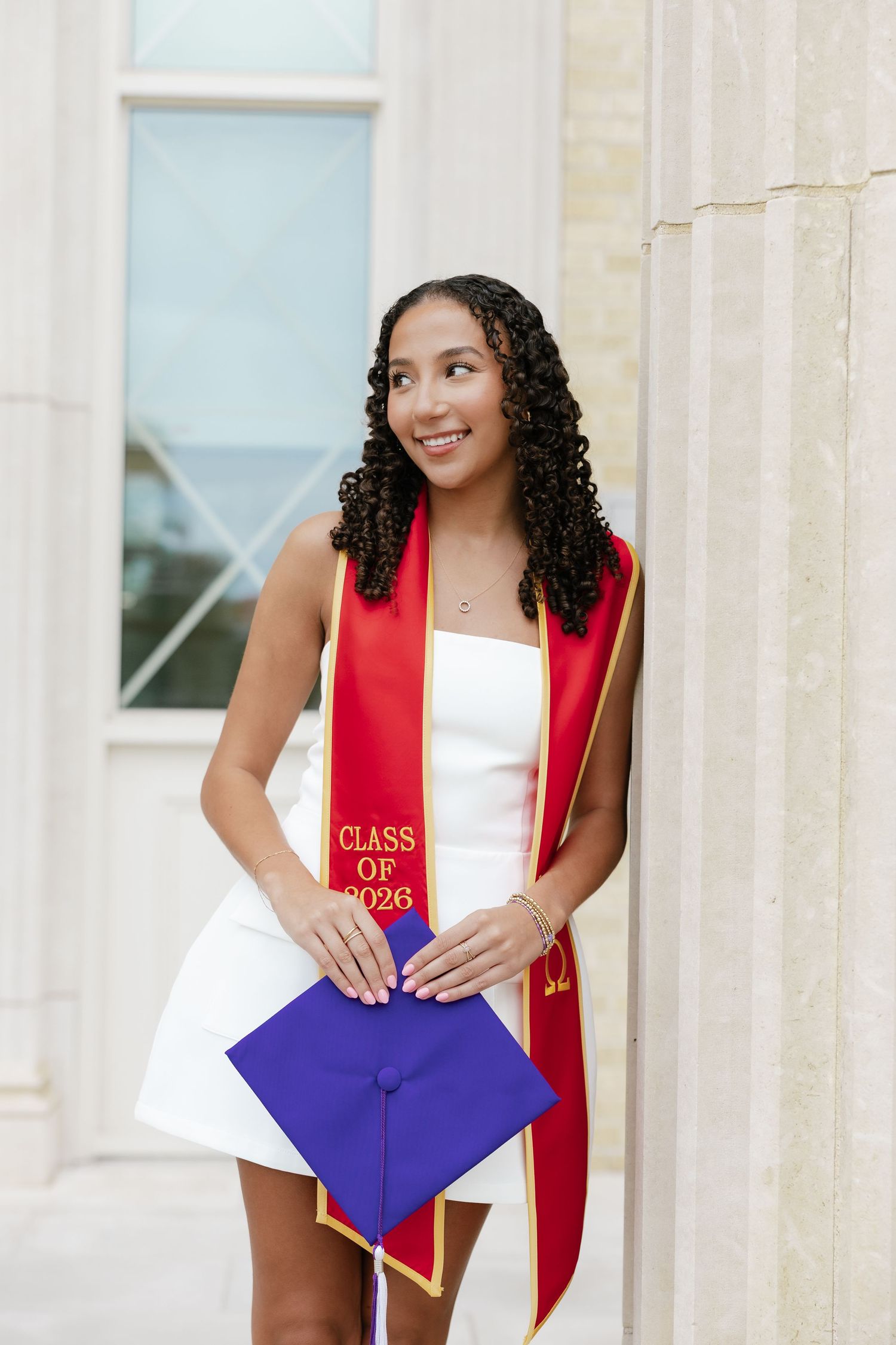 Smiling graduate in white dress with red Class of 2026 stole holds blue cap by stone columns.