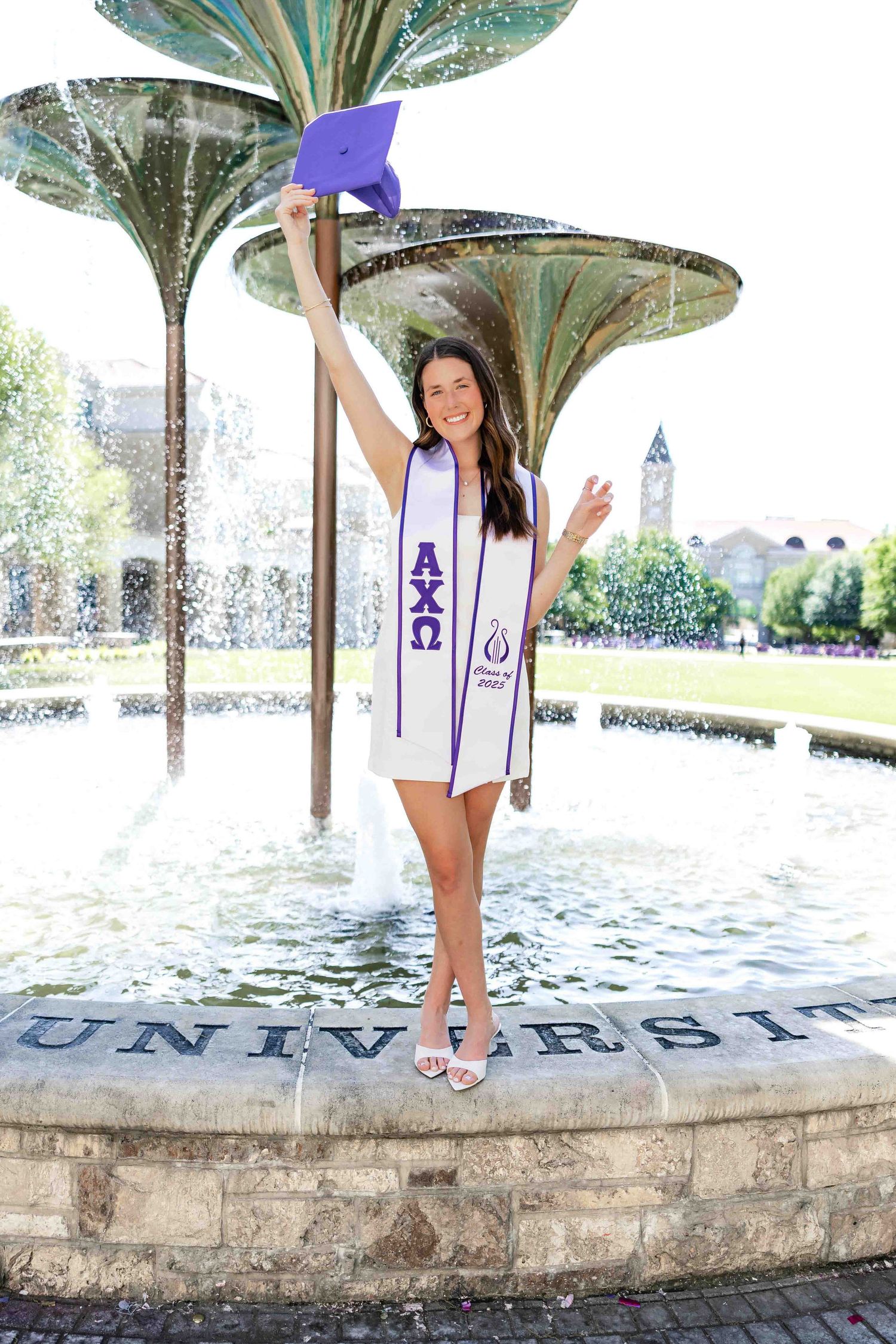 Graduate in white graduation stole celebrating in campus fountain.