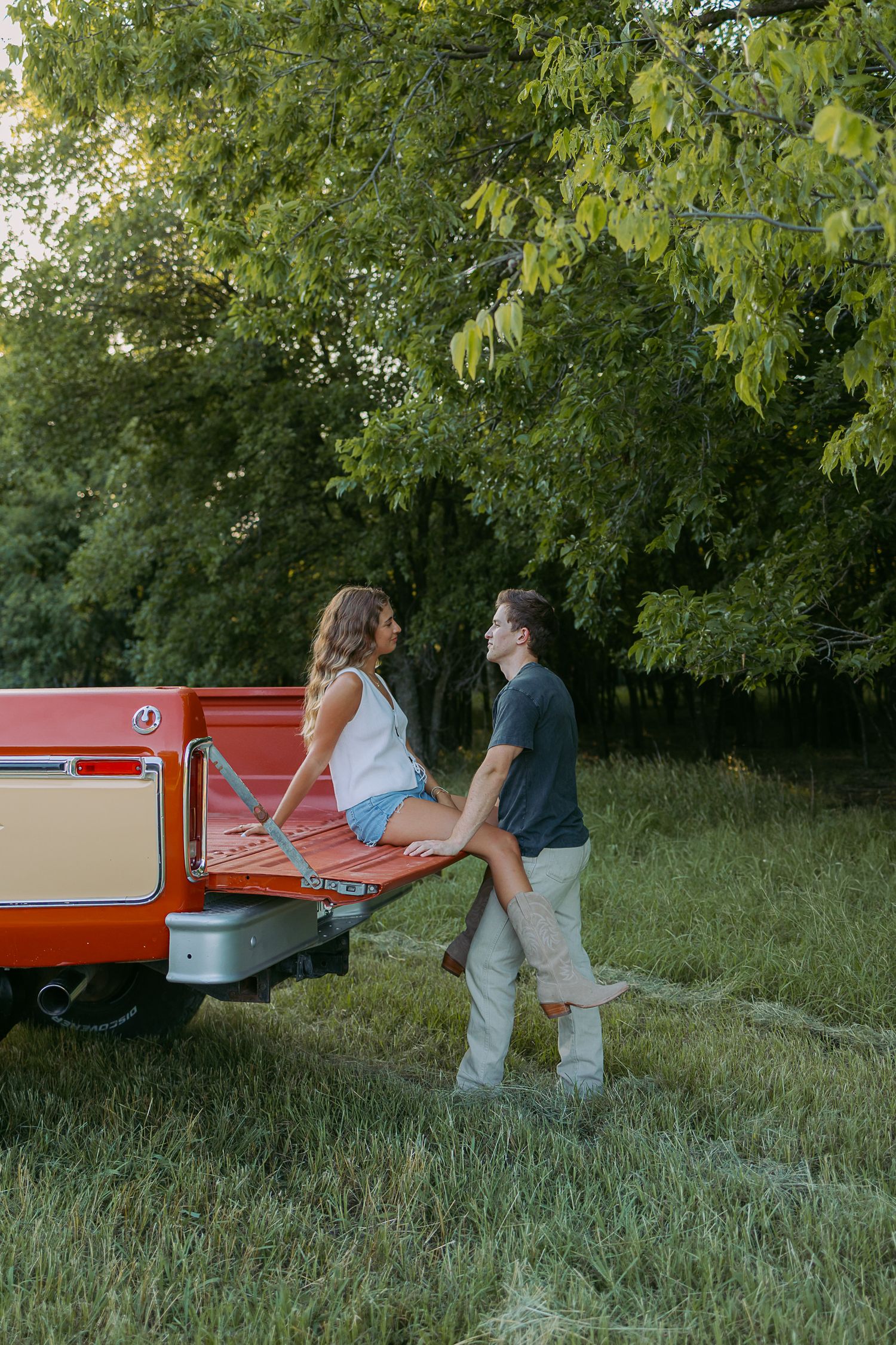 A couple sits together on the tailgate of a vintage red pickup truck in a grassy field surrounded by trees.