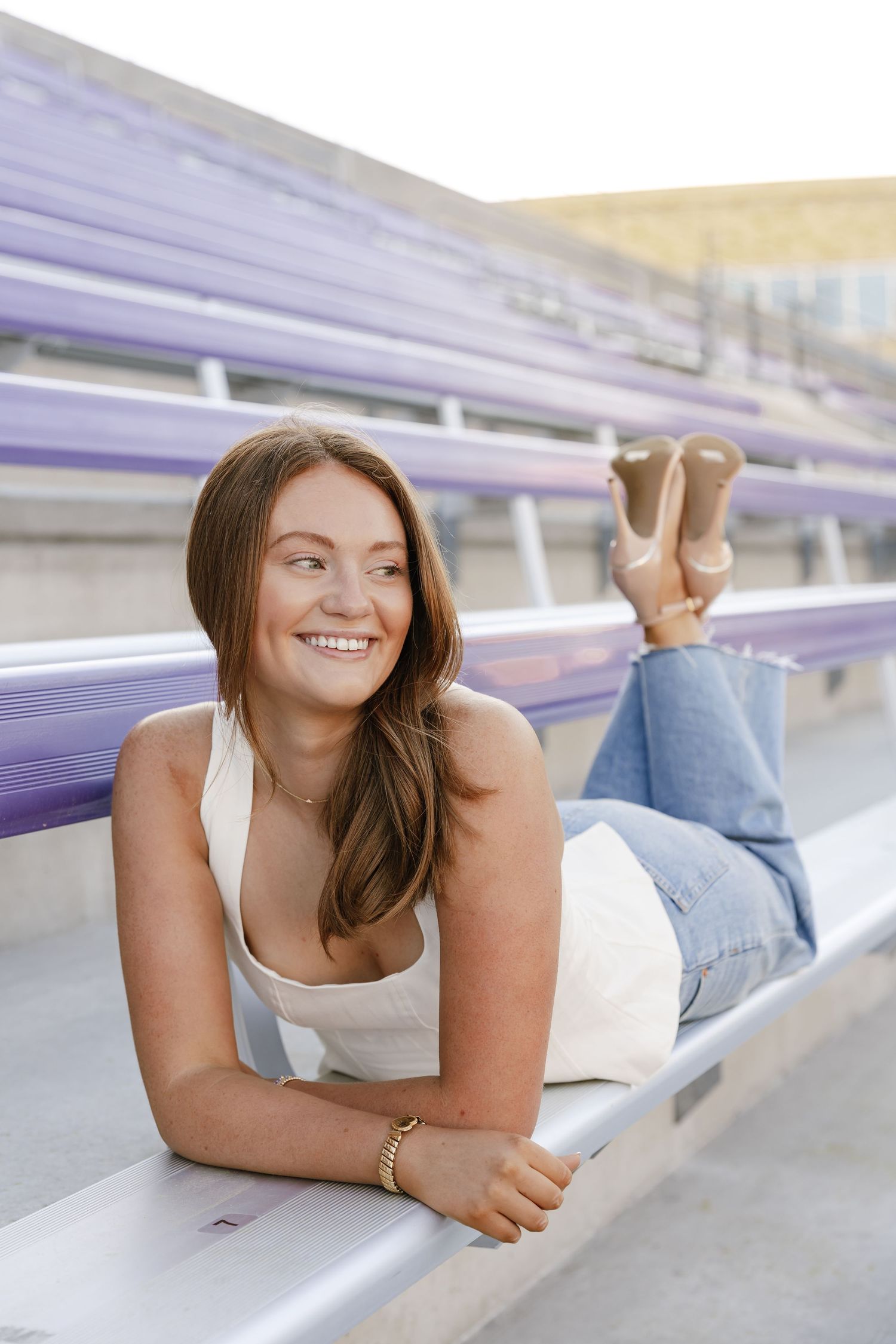 Smiling young woman in white top and jeans lying on purple stadium bleachers, looking away cheerfully.