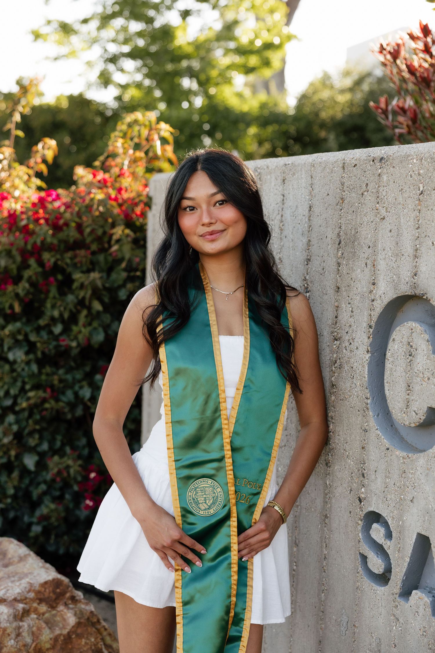 Young woman in white dress wearing green and gold graduation sash poses proudly outdoors near stone monument.