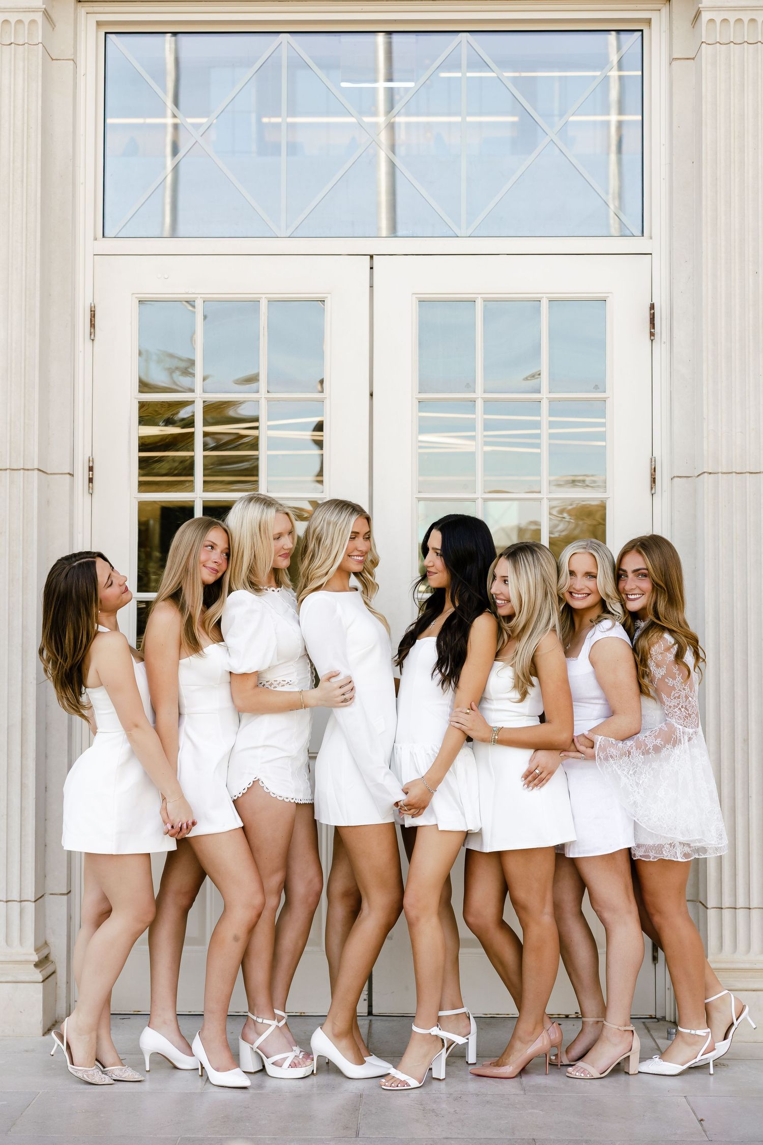 Eight graduates in white dresses and heels pose together smiling outside Chapman University Doors.