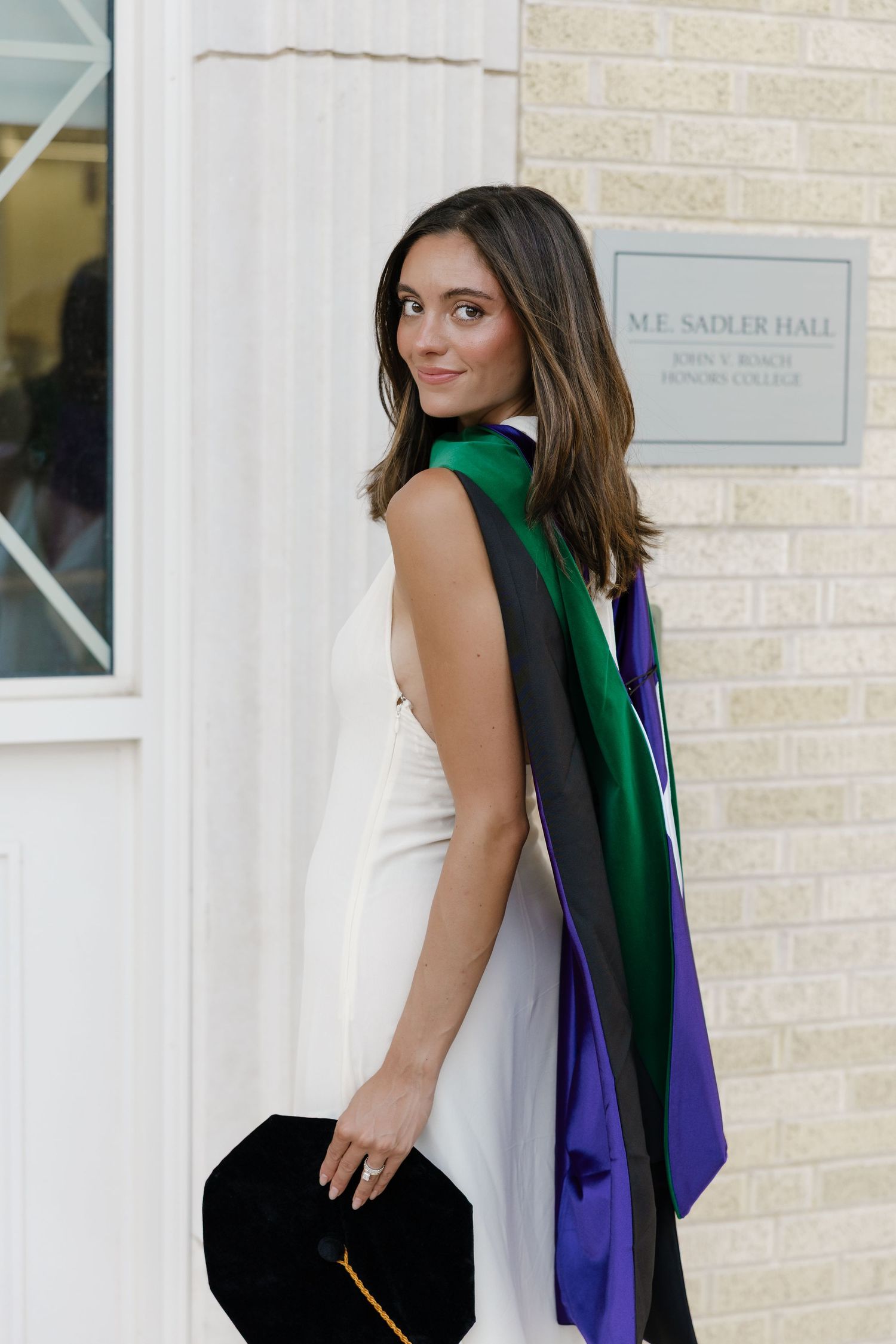 Young woman in white dress and graduation regalia smiles over shoulder near M.E. Sadler Hall Honors College sign.