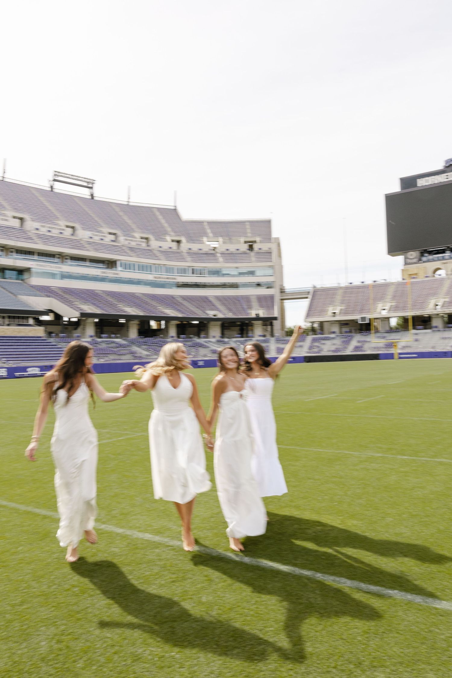 Four women in white dresses walk hand-in-hand across a football stadium field, celebrating joyfully.