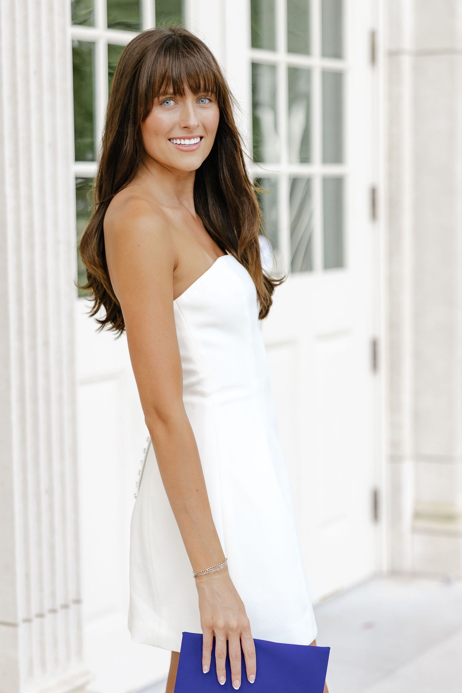 A smiling brunette woman in a white strapless dress holds a blue clutch purse while posing outdoors.