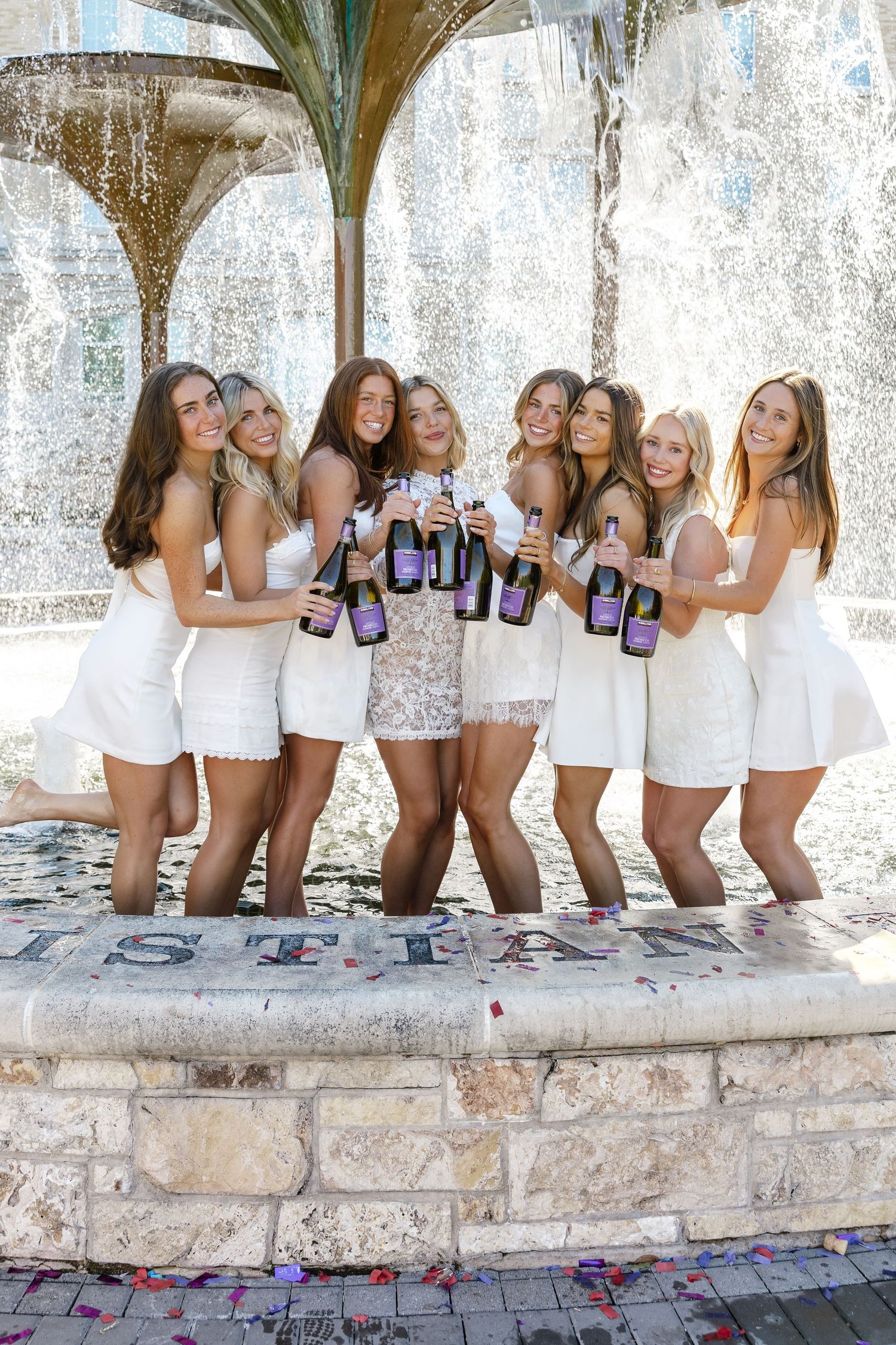 Group of women in white dresses celebrating with champagne bottles by a fountain, tossing confetti.