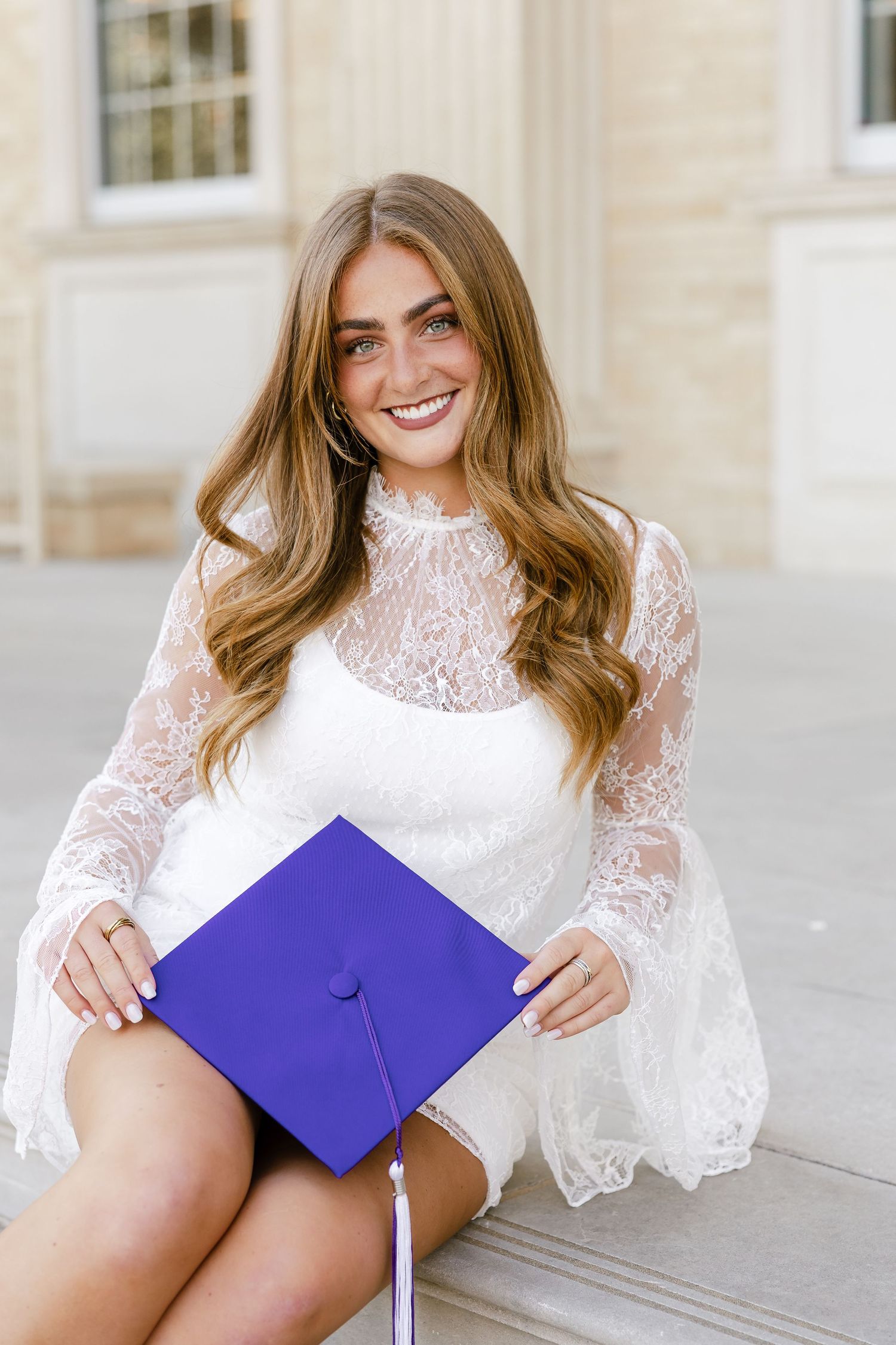 Smiling young woman in white lace dress holding purple graduation cap, seated outdoors near a stone building.