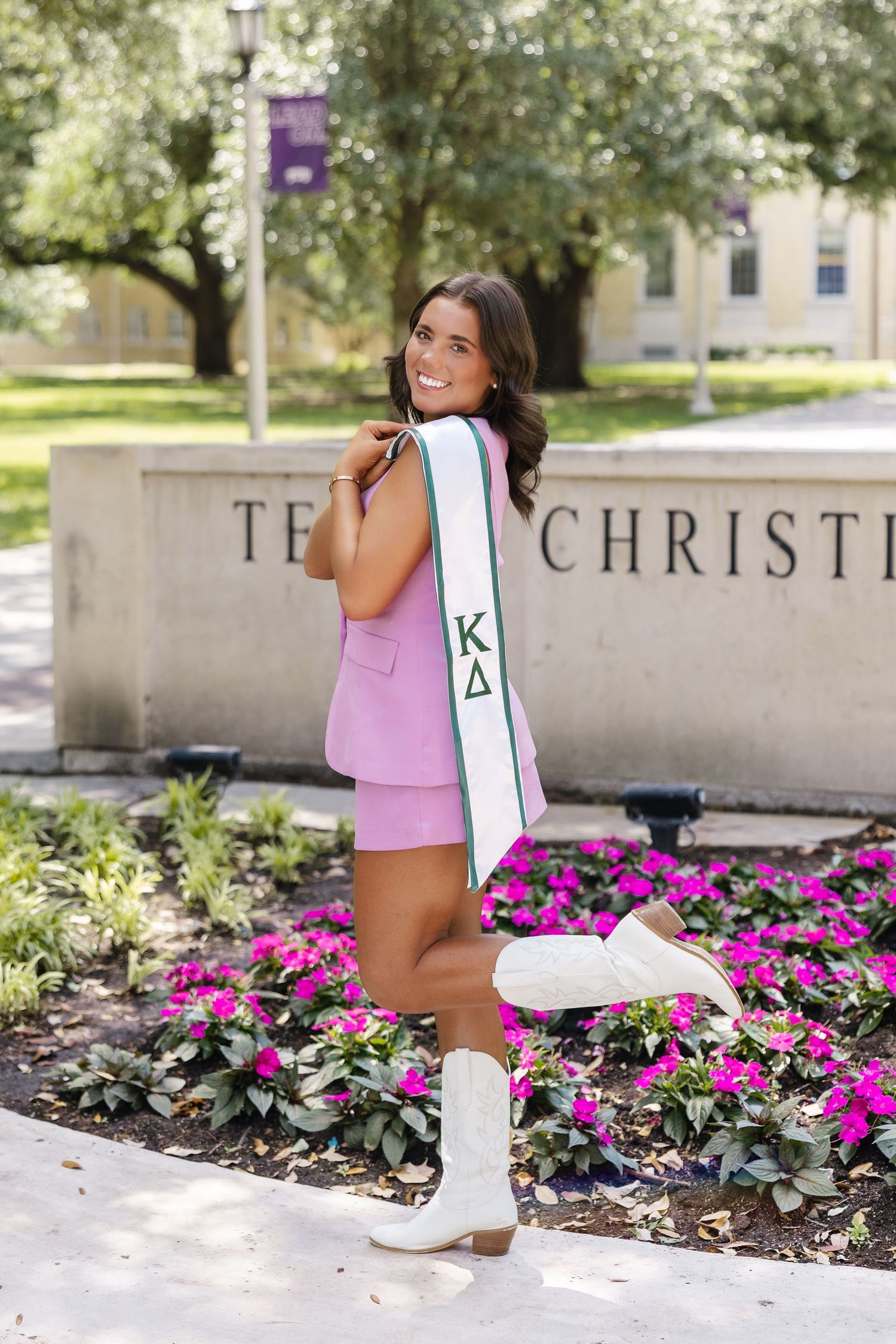 Young woman in pink outfit wearing Kappa Delta sorority sash poses at Texas Christian University campus sign.