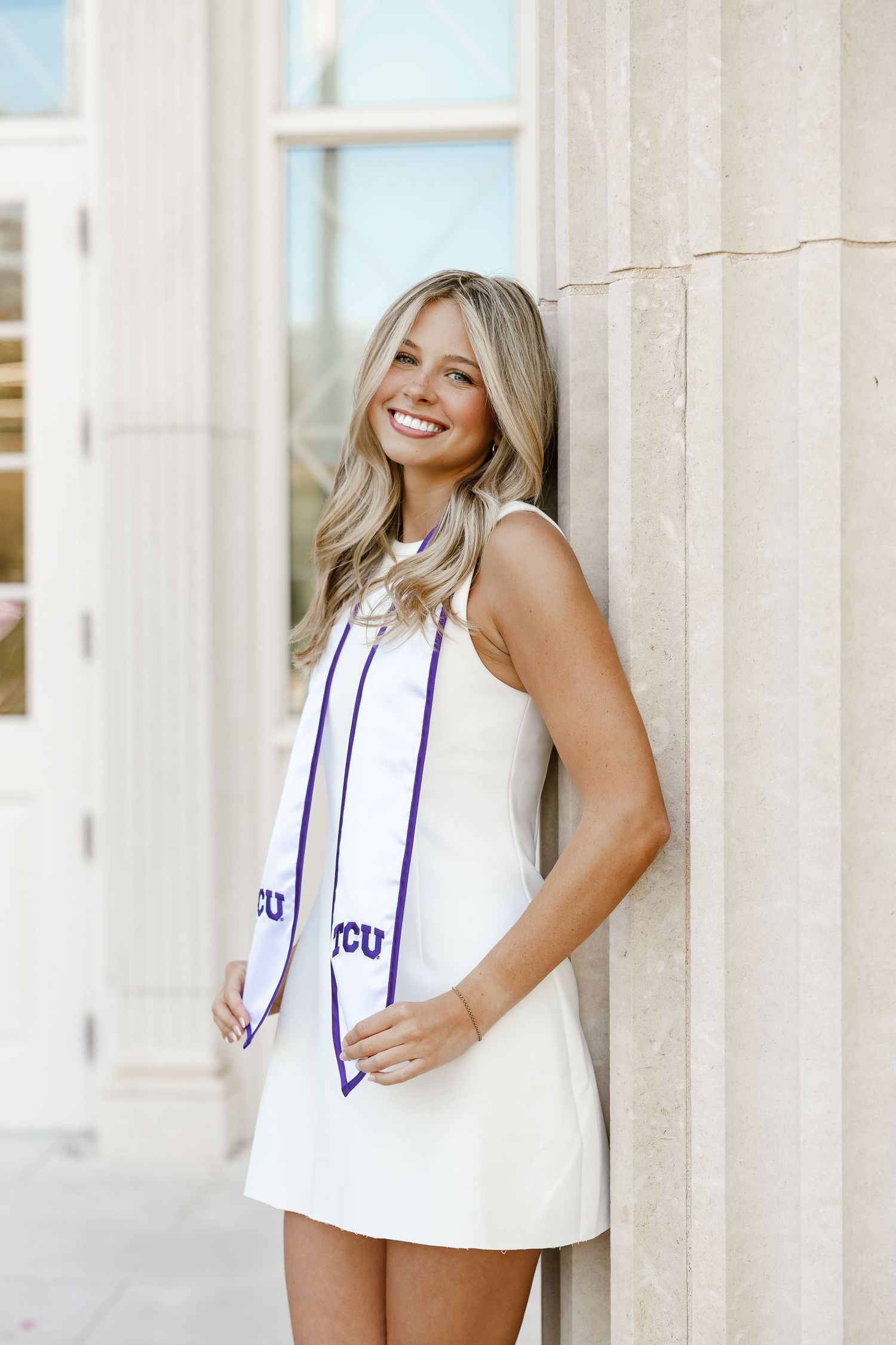 Smiling blonde woman in white dress and TCU graduation stole leaning against a column outdoors.