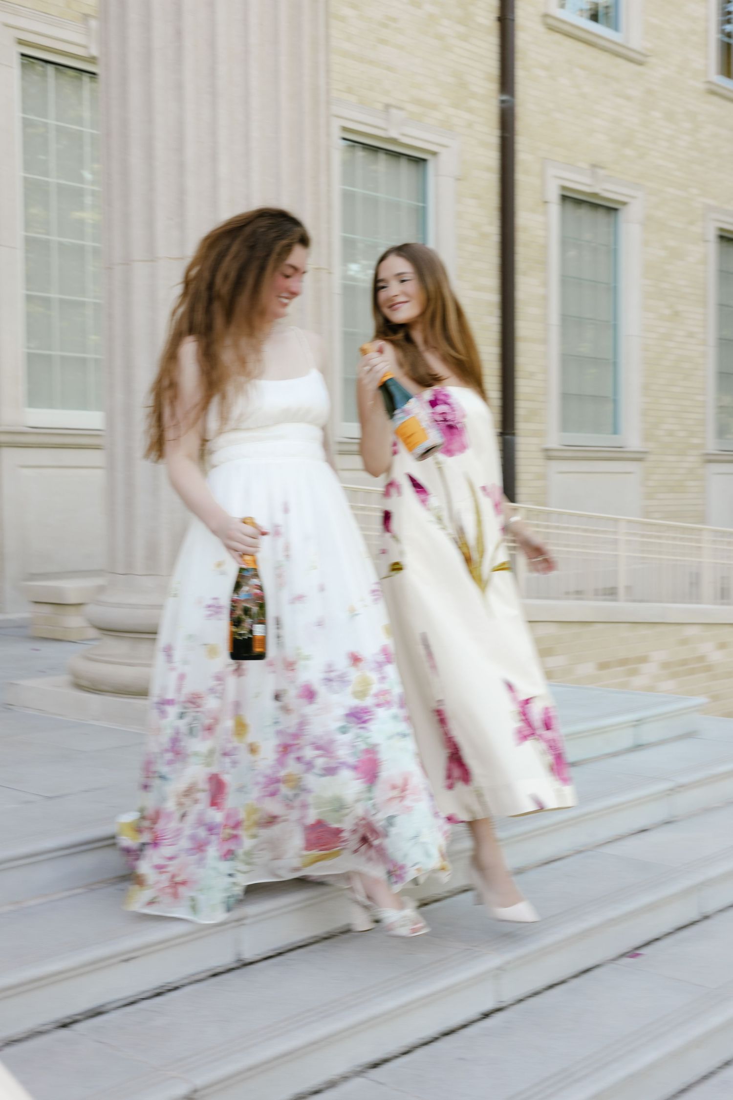 Two women in floral maxi dresses descend stone steps holding champagne bottles outdoors.