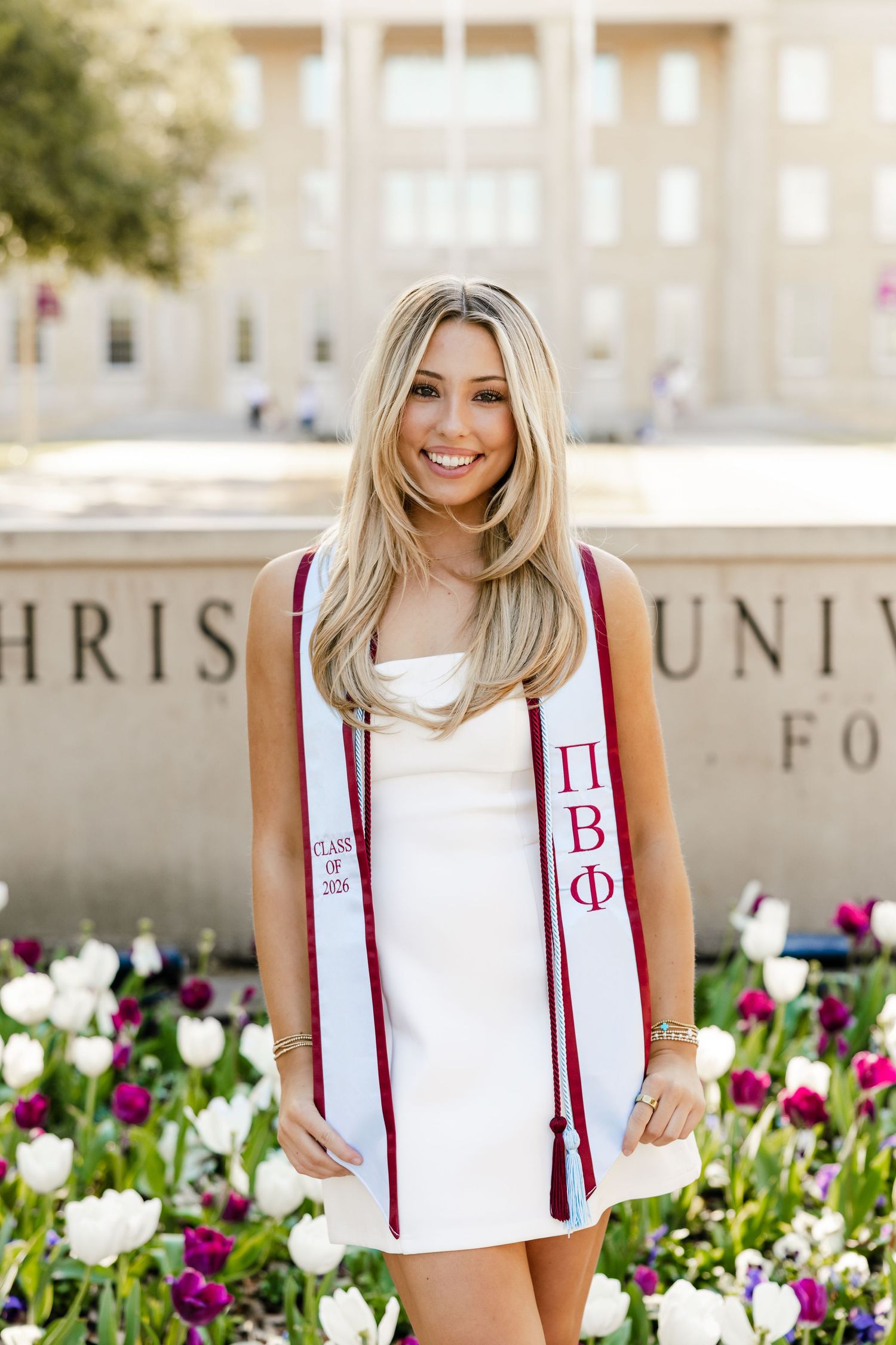 Smiling blonde woman in white dress wearing Pi Beta Phi graduation stole at TCU.