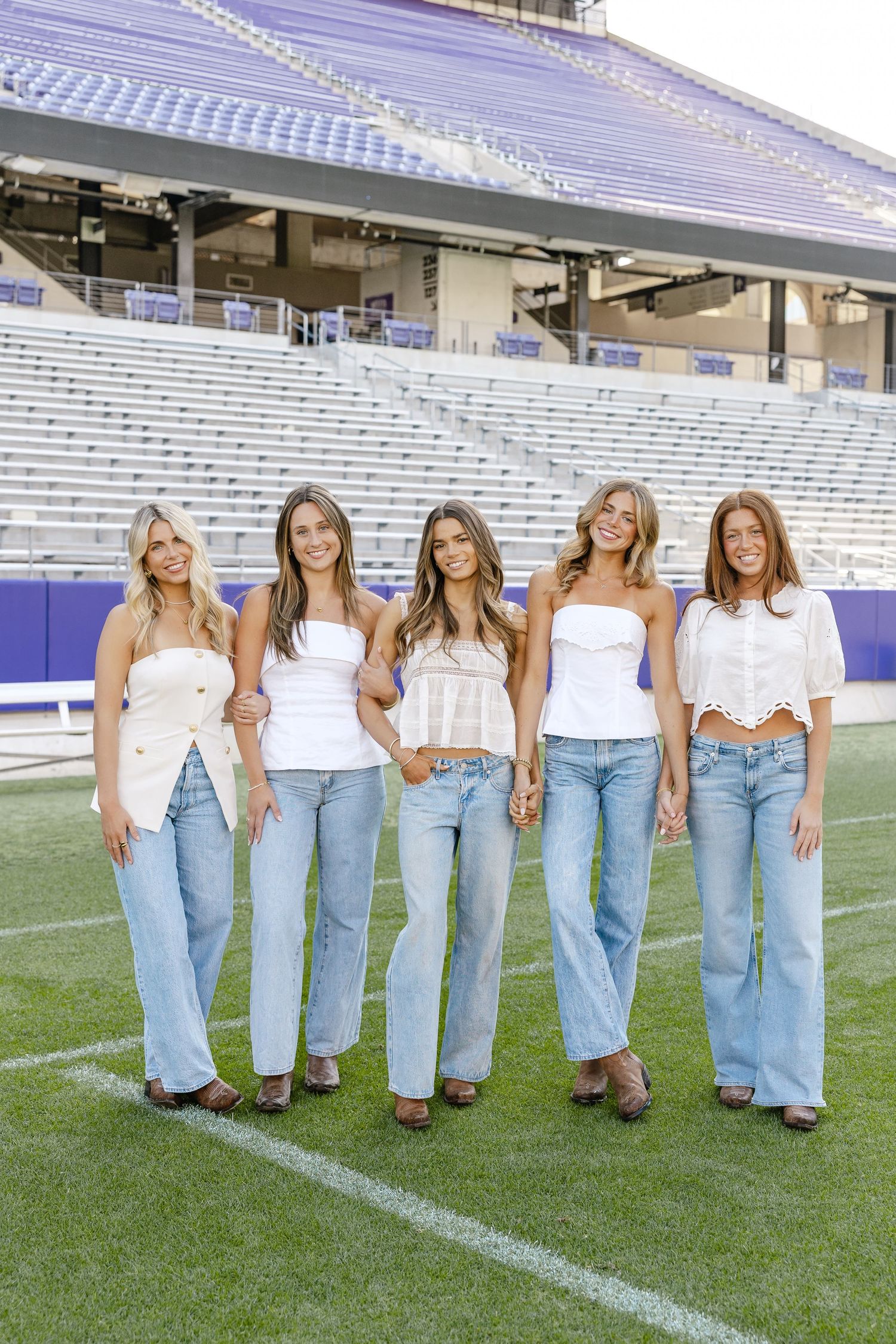 Five young women in white tops and light blue jeans pose together on a football field stadium.