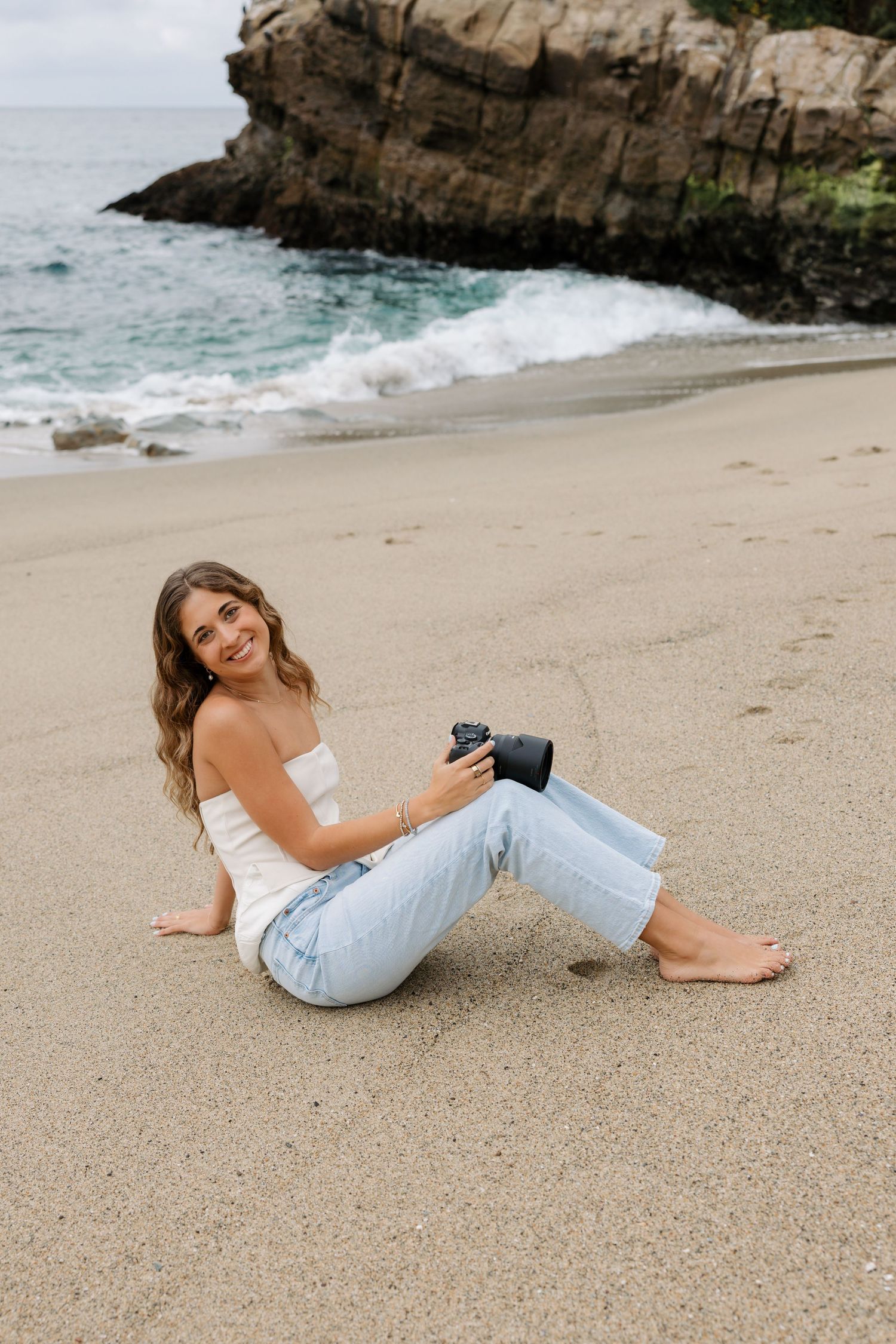 Person in white top and light jeans relaxes on sandy beach near rocky coastal cliffs and ocean waves.