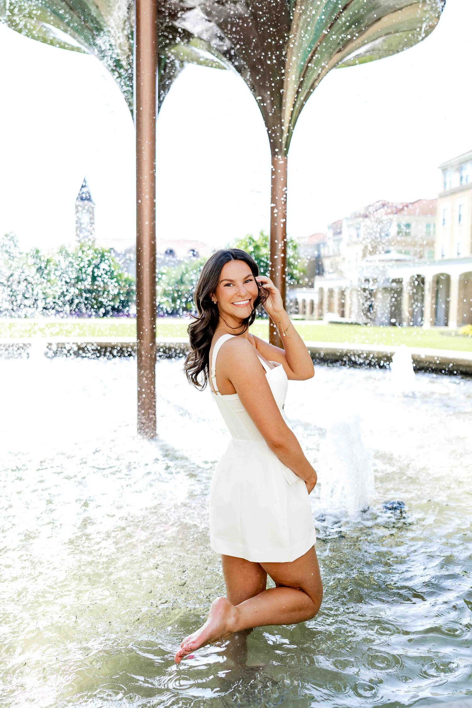 Person in white dress poses playfully by decorative fountain with palm tree sculptures.
TCU Graduation photographer