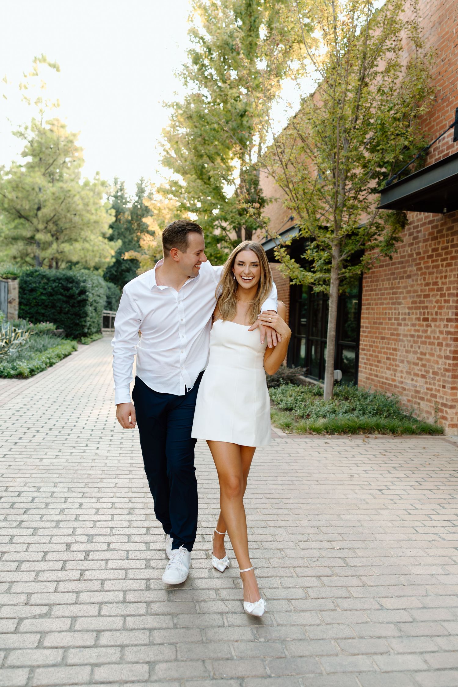 A romantic couple playfully walks together on a brick pathway surrounded by green trees.
