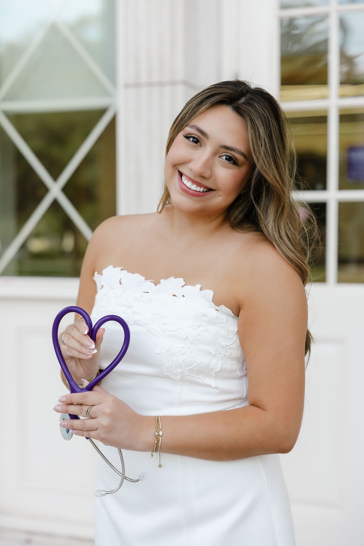 Smiling young woman in white strapless dress holding a purple heart-shaped stethoscope, posing outdoors at Chapman University