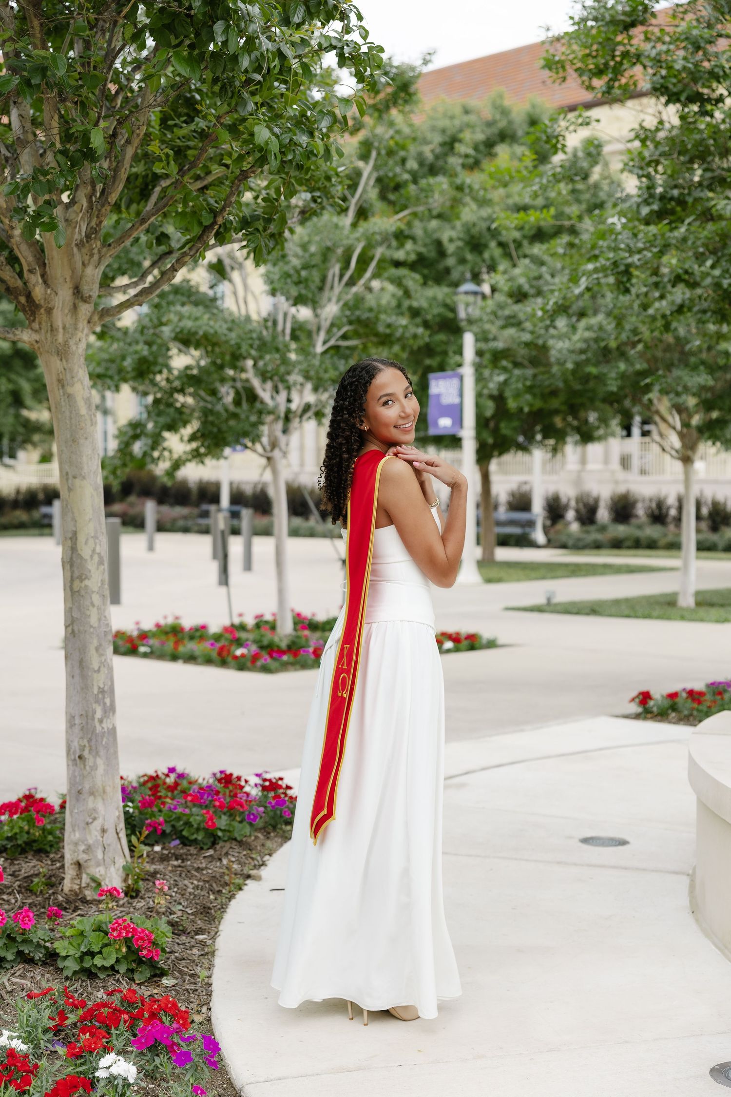 Young woman in white gown and red graduation sash poses outdoors near colorful flowers on a university campus.
