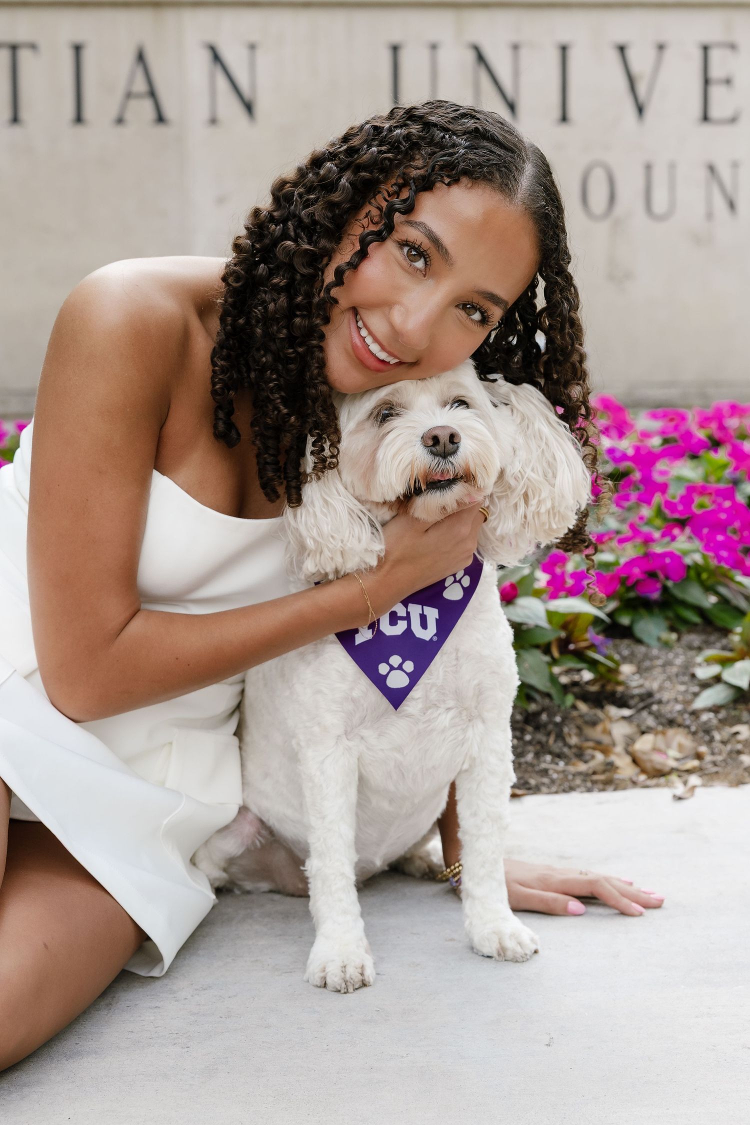 A smiling woman in a white dress hugs a fluffy white dog wearing a purple TCU bandana near a university building.