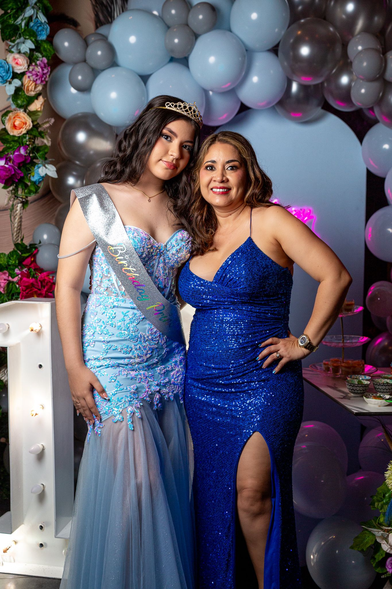 Two people in sparkly formal dresses pose together against a balloon arch backdrop with floral decorations.