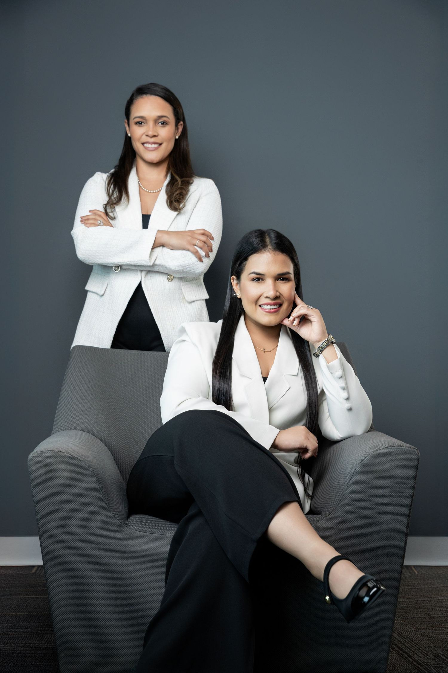Two professional women in white blazers pose confidently in a modern office setting with gray walls.