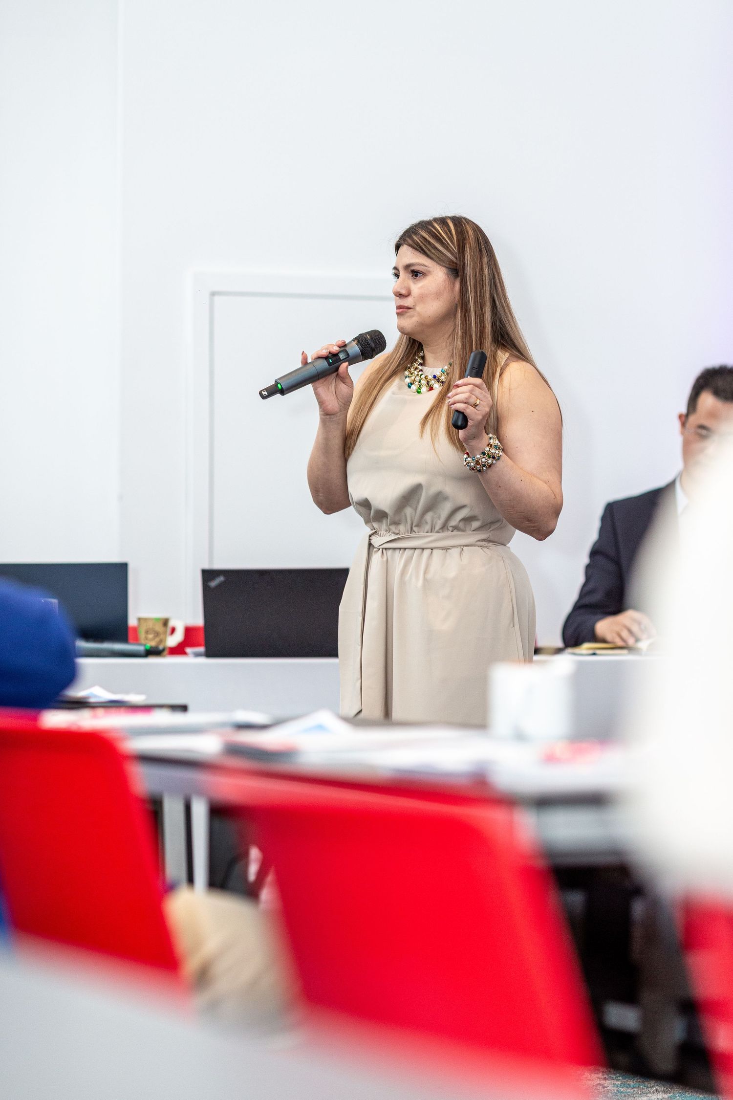 A speaker in a beige dress presents at a conference room with red chairs visible in the foreground.