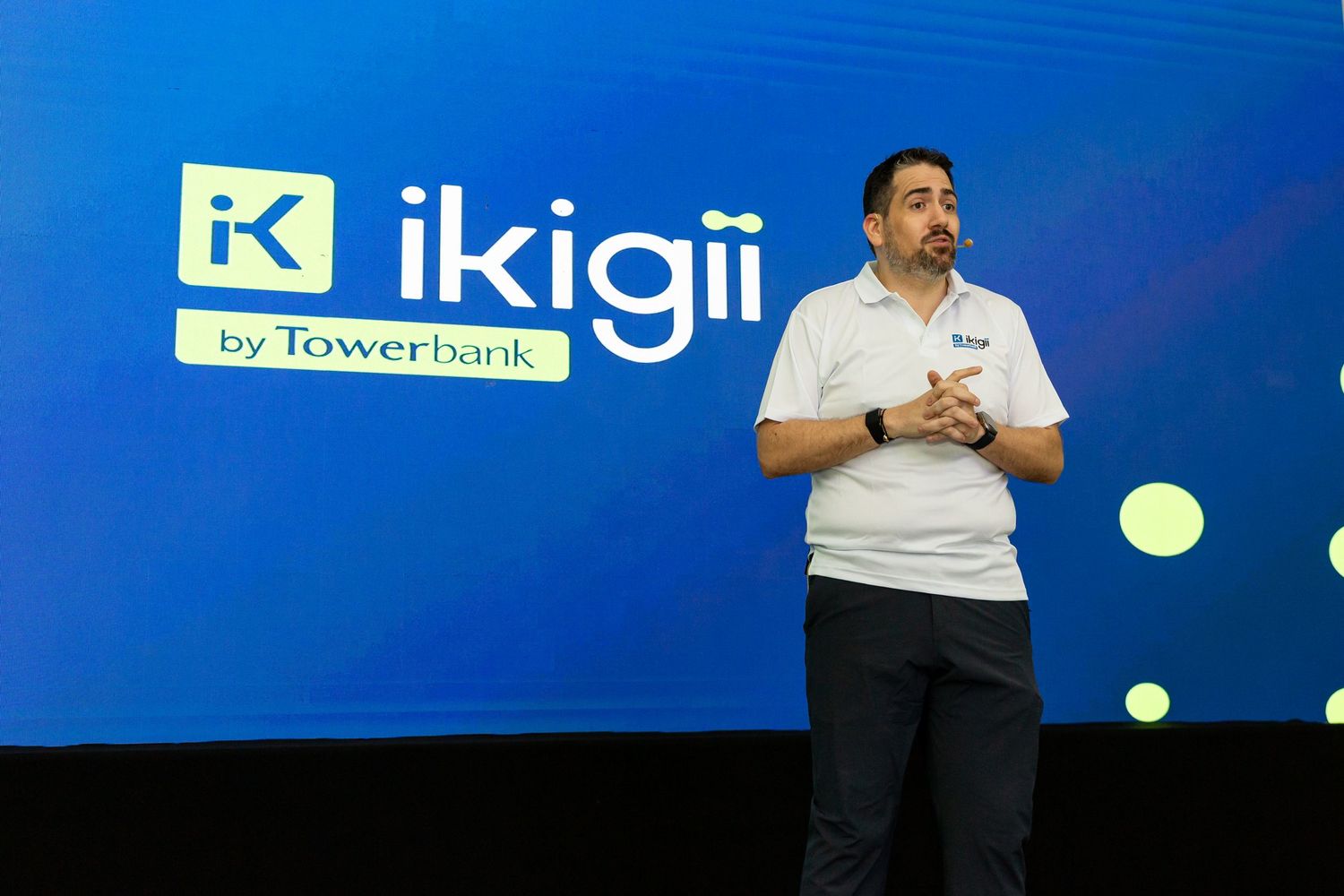 A man wearing a white shirt stands in front of a large blue background with the Ikigii logo.