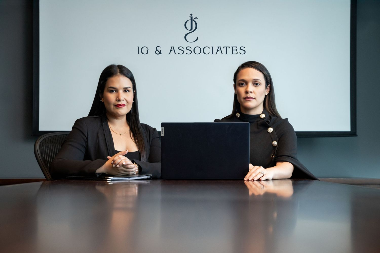Two professional women sit at a conference table with a laptop in front of an IG & Associates sign.