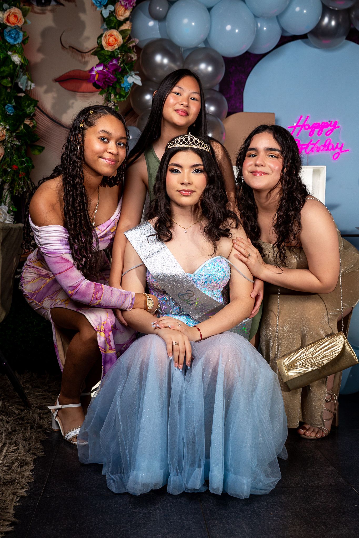 A celebration photo with a blue tulle dress and tiara against a balloon arch and floral backdrop at a birthday party.
