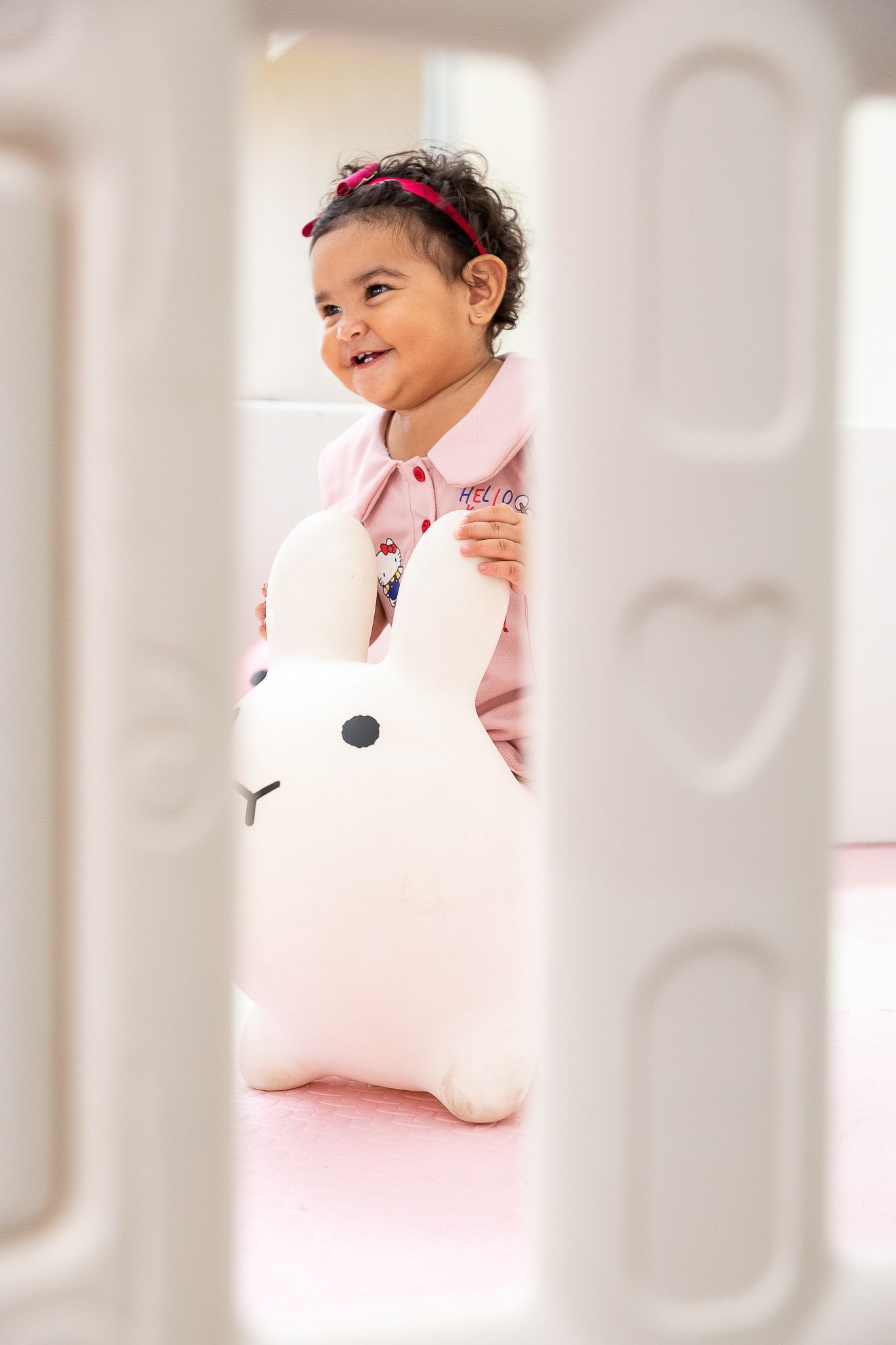Smiling baby girl in pink hoodie holding a white bunny night light toy inside a white crib.