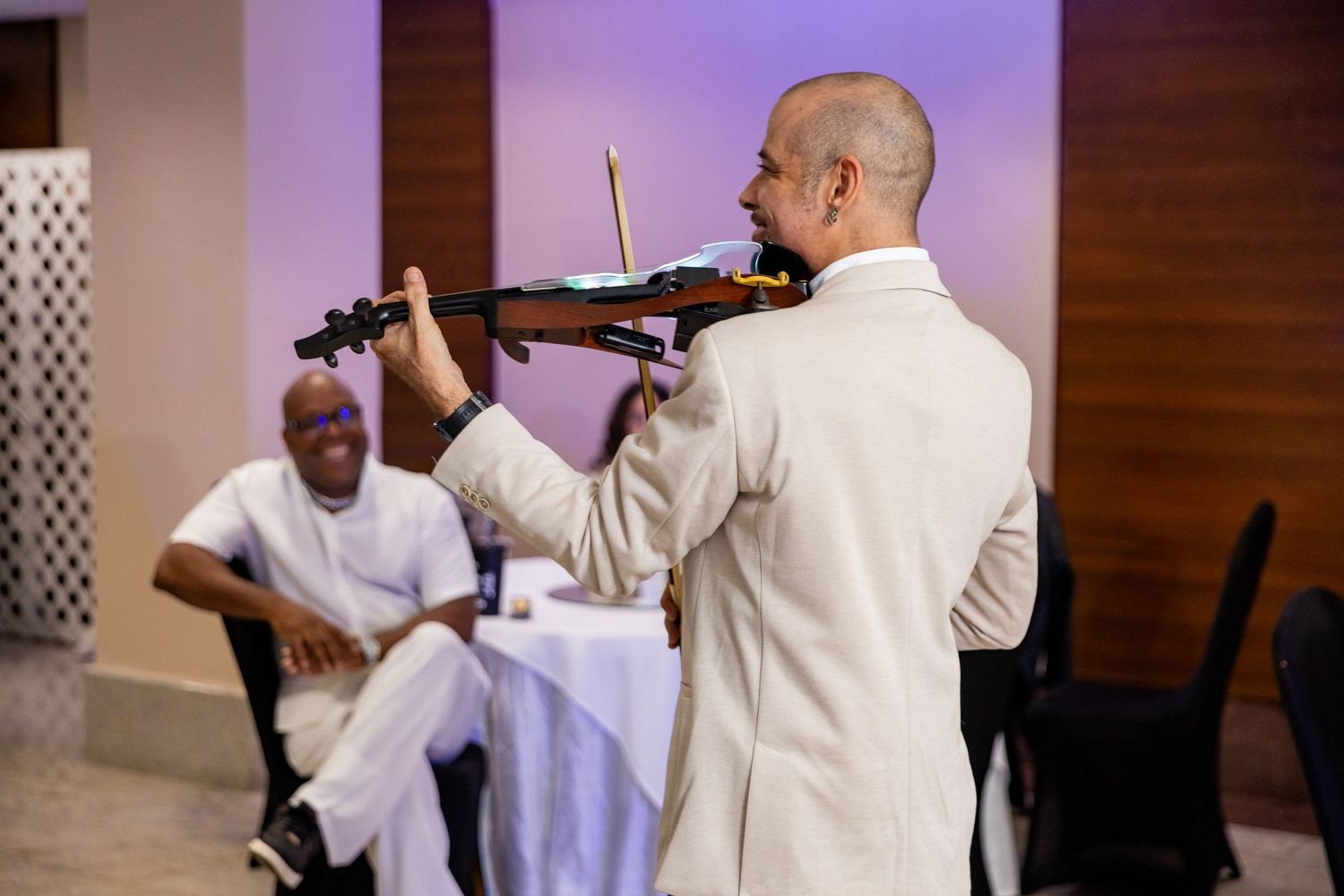 Violinist in white suit performs for smiling guest at elegant indoor event.