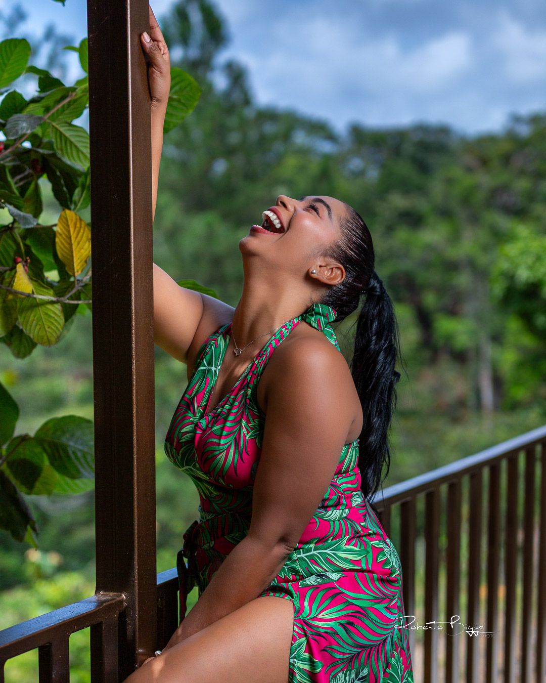 Woman in tropical print dress poses expressively on outdoor balcony with lush green forest backdrop in natural lighting.