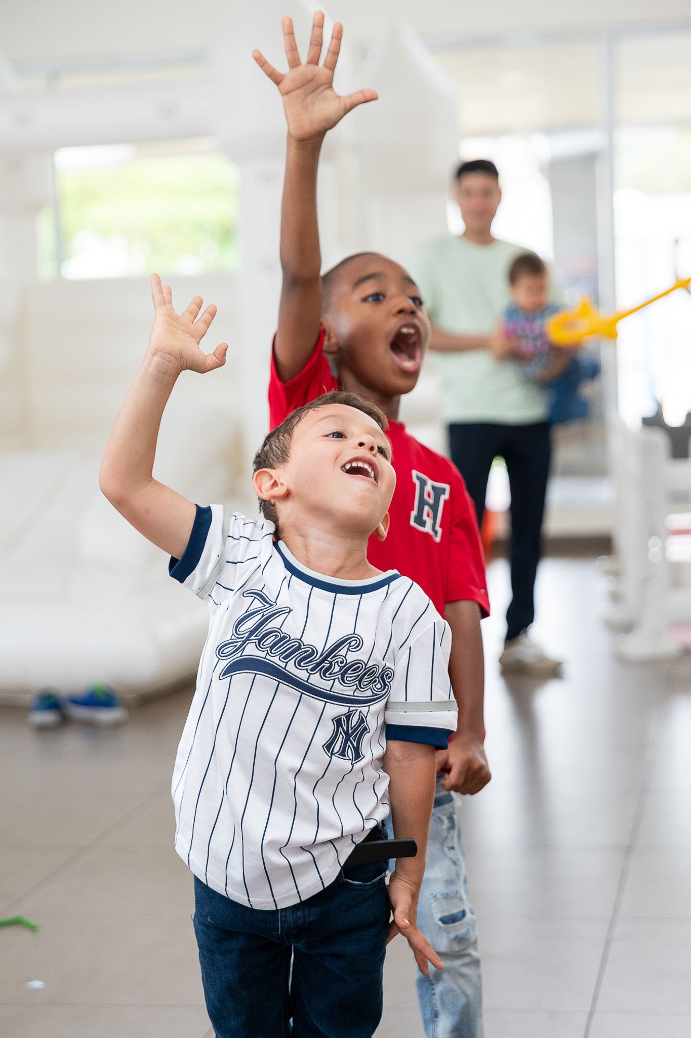 Two excited boys reaching up with hands raised, wearing Yankees jersey and red shirt, playing indoors.
