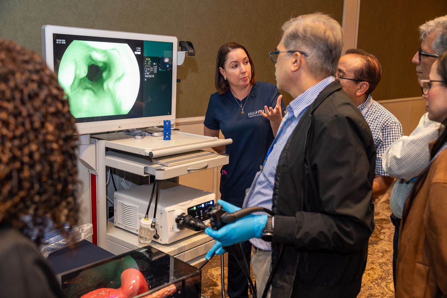 Medical professionals discuss endoscopy simulation technology at a conference, viewing colonoscopy imaging on a monitor.