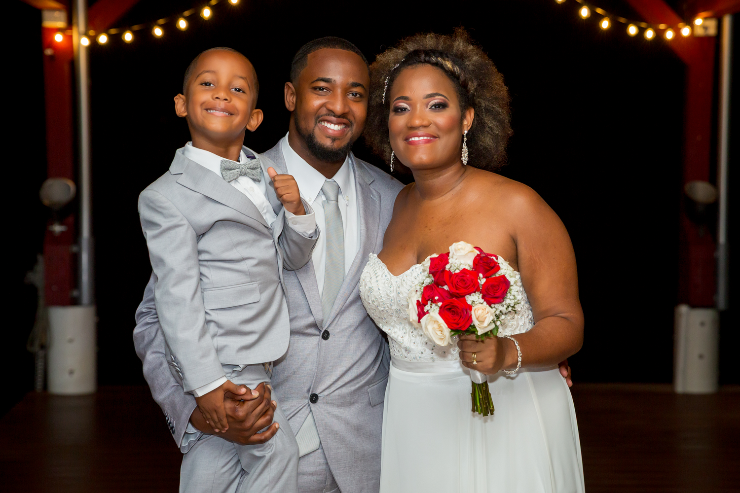 A happy wedding portrait of three people dressed in white against a dark background with string lights.