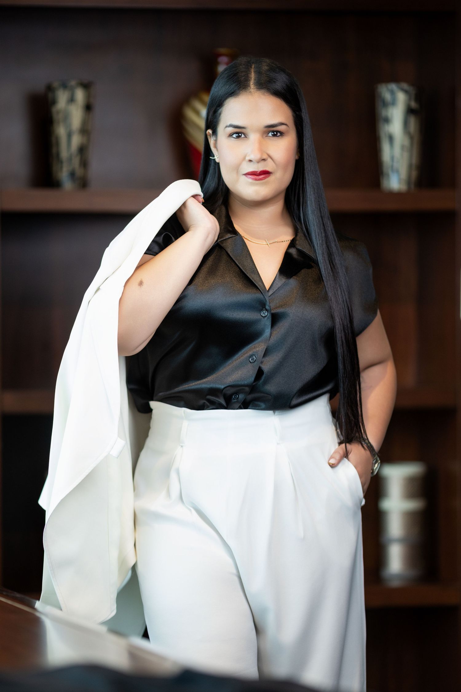 A confident businesswoman in black satin blouse and white trousers holds a blazer over her shoulder in an office setting.
