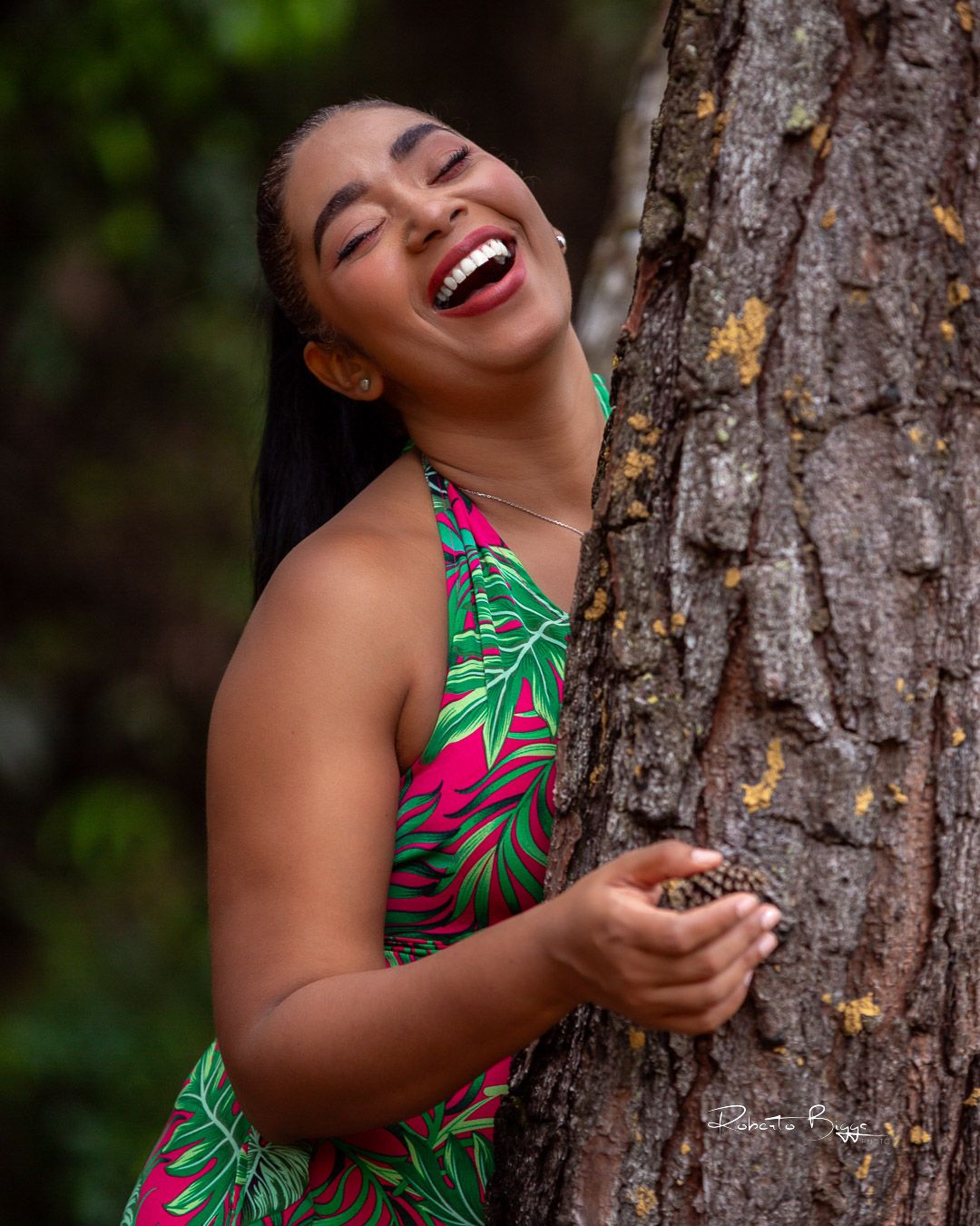 A joyful expression lights up the photo as someone in a tropical print dress leans against a tree trunk outdoors.