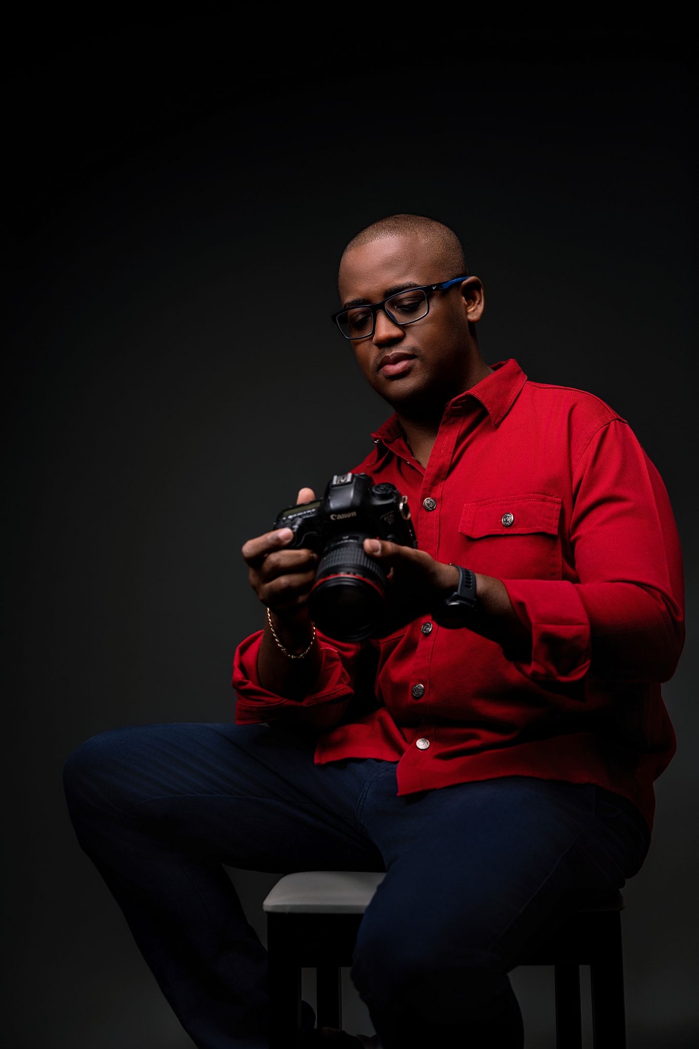 A photographer in a red shirt and dark pants sits in dramatic lighting while examining a camera against a black background.