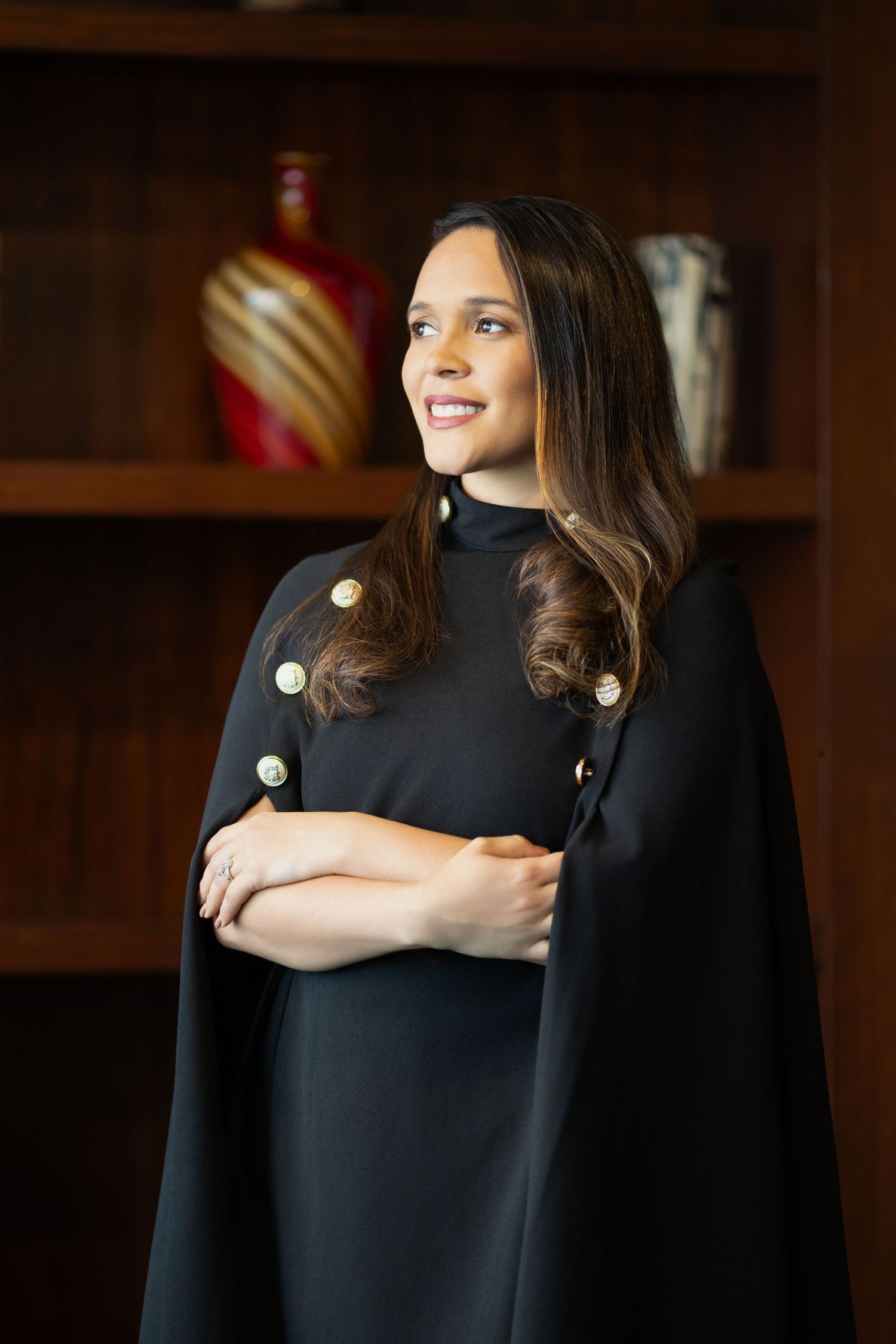 Professional woman in elegant black cape dress with gold buttons, arms crossed, smiling confidently.